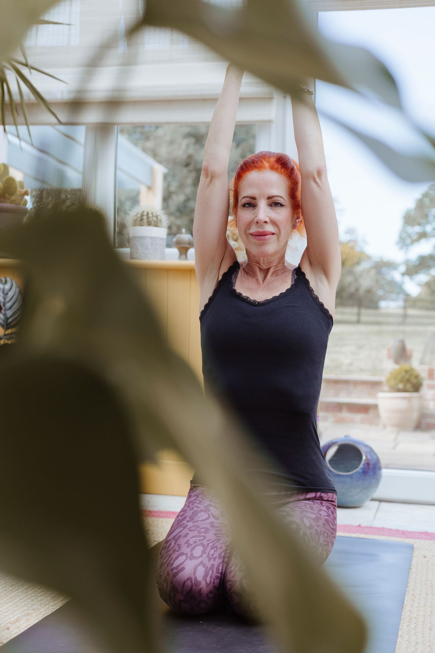 A woman with red hair kneels on a yoga mat indoors, arms raised overhead in a calm, focused pose, partly framed by a large plant in the fore