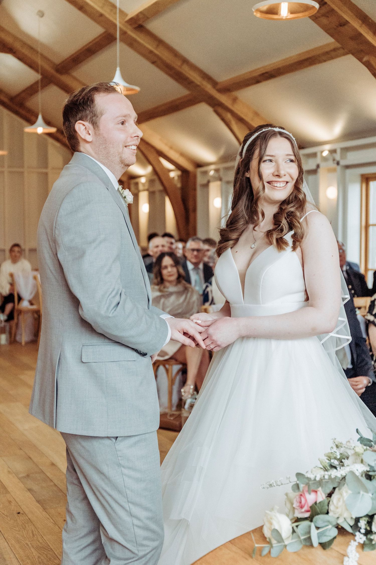 A bride and groom hold hands and smile during their ceremony, surrounded by guests in a warm, light-filled venue.