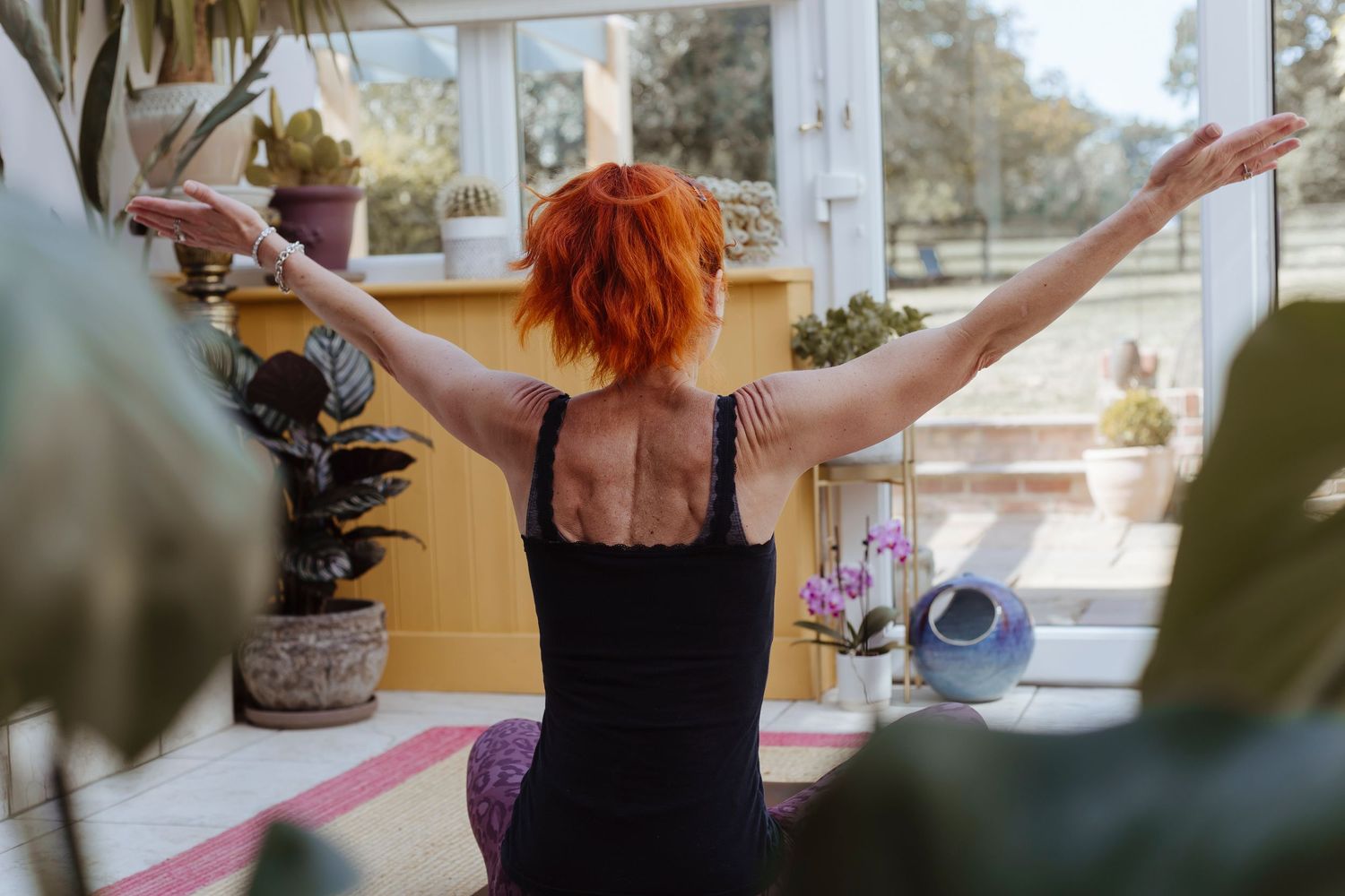 A woman with red hair sits on a mat, arms extended wide in a calm, open yoga pose inside a bright, plant-filled room.