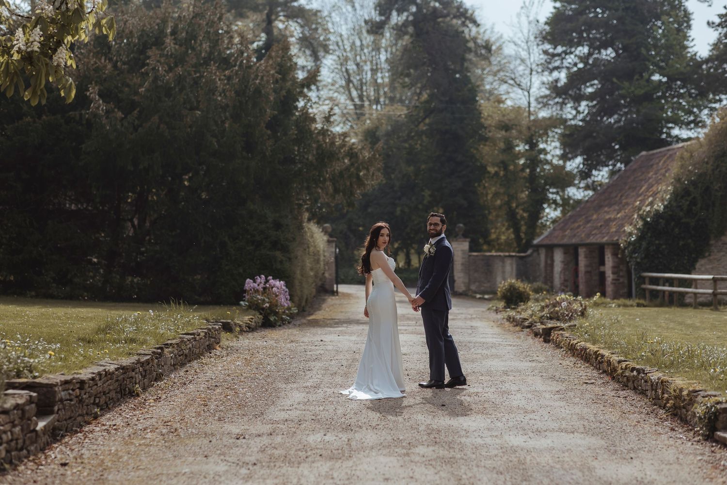 A couple in wedding attire stands hand in hand on a quiet, tree-lined path beside rustic buildings.