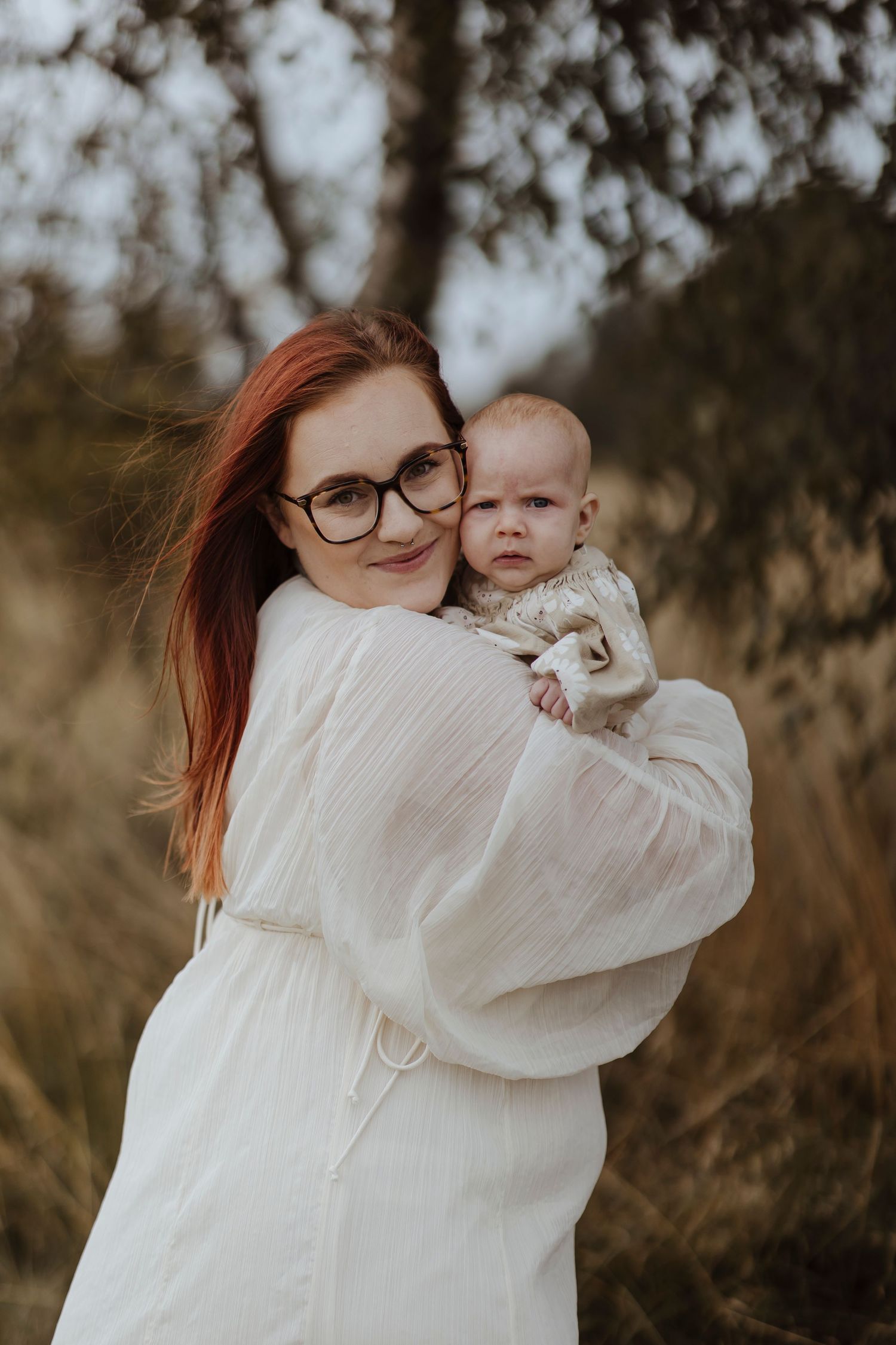 A woman in glasses holds a baby outdoors, both looking toward the camera with a calm, gentle expression.