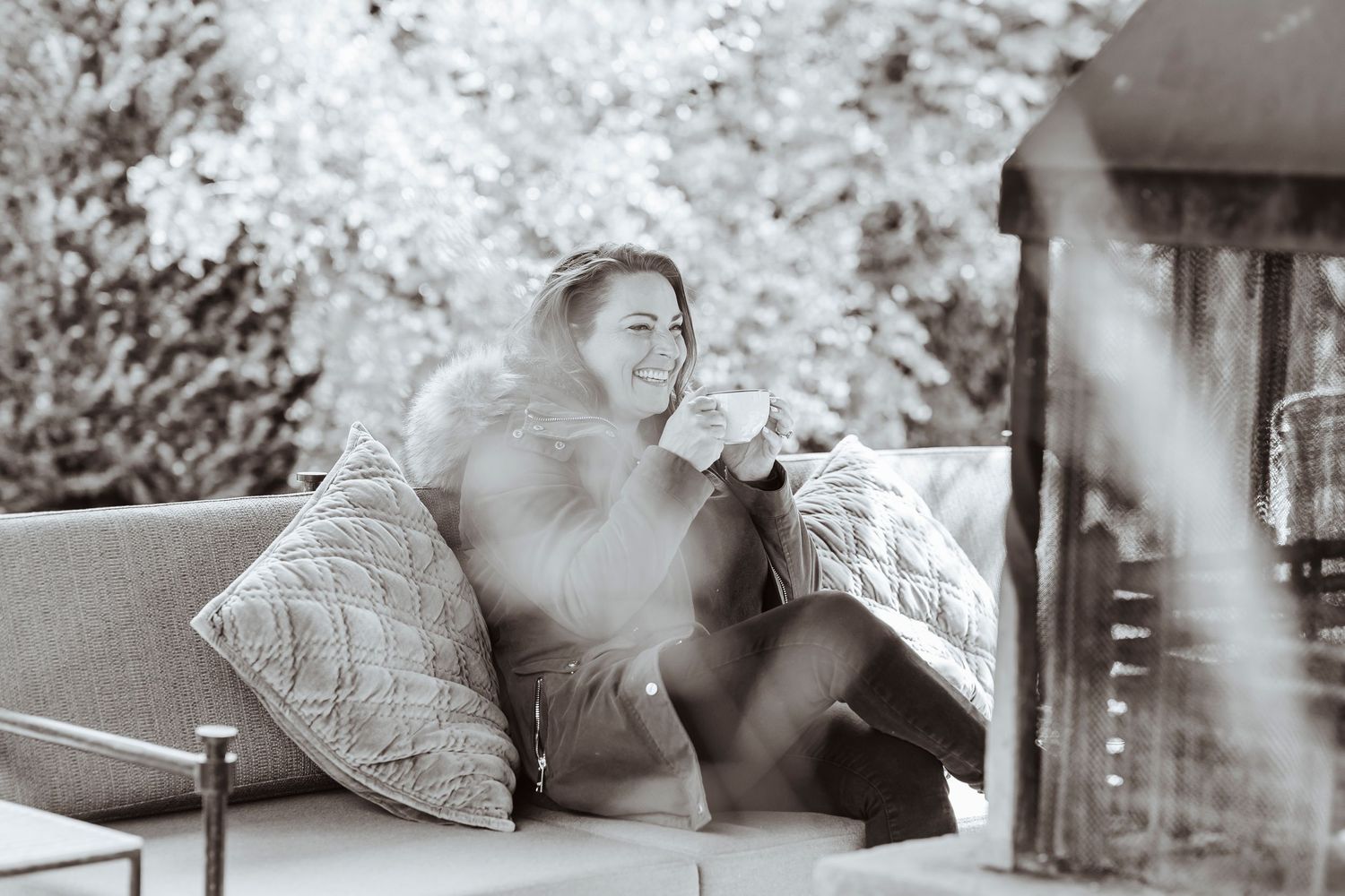 A person sits bundled up on an outdoor sofa, smiling as they enjoy a warm drink in a peaceful, cozy setting.