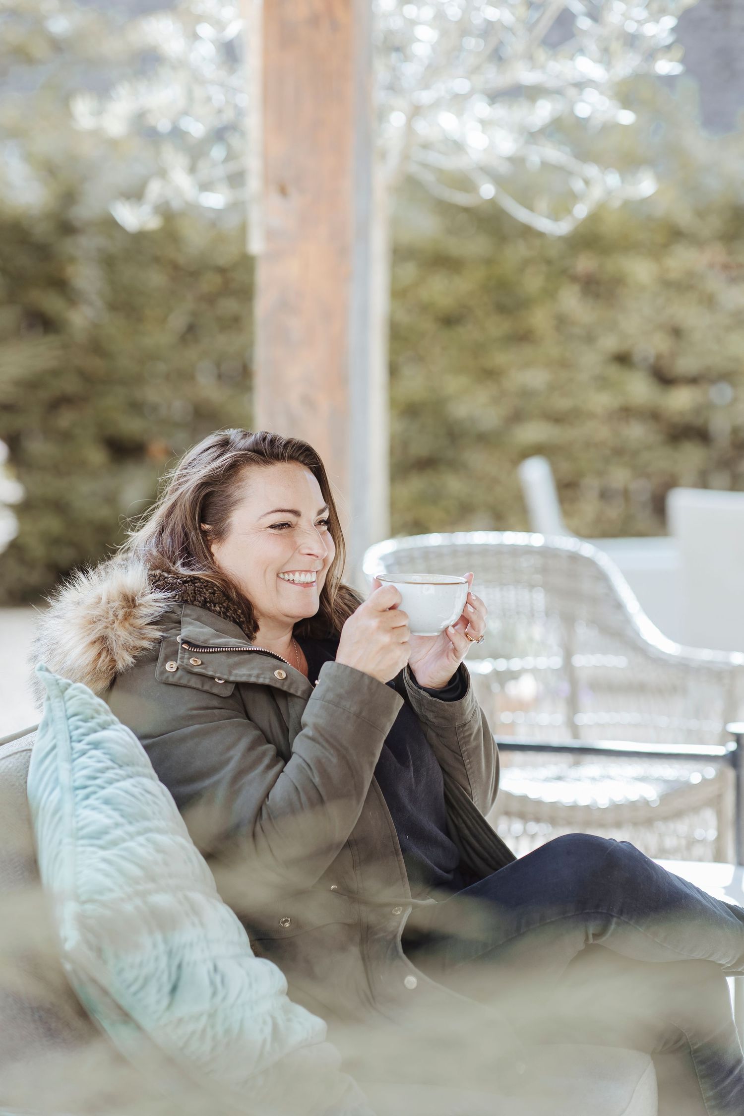 A woman relaxes outdoors with a warm drink, wrapped in a cozy coat and smiling in the soft, bright light.