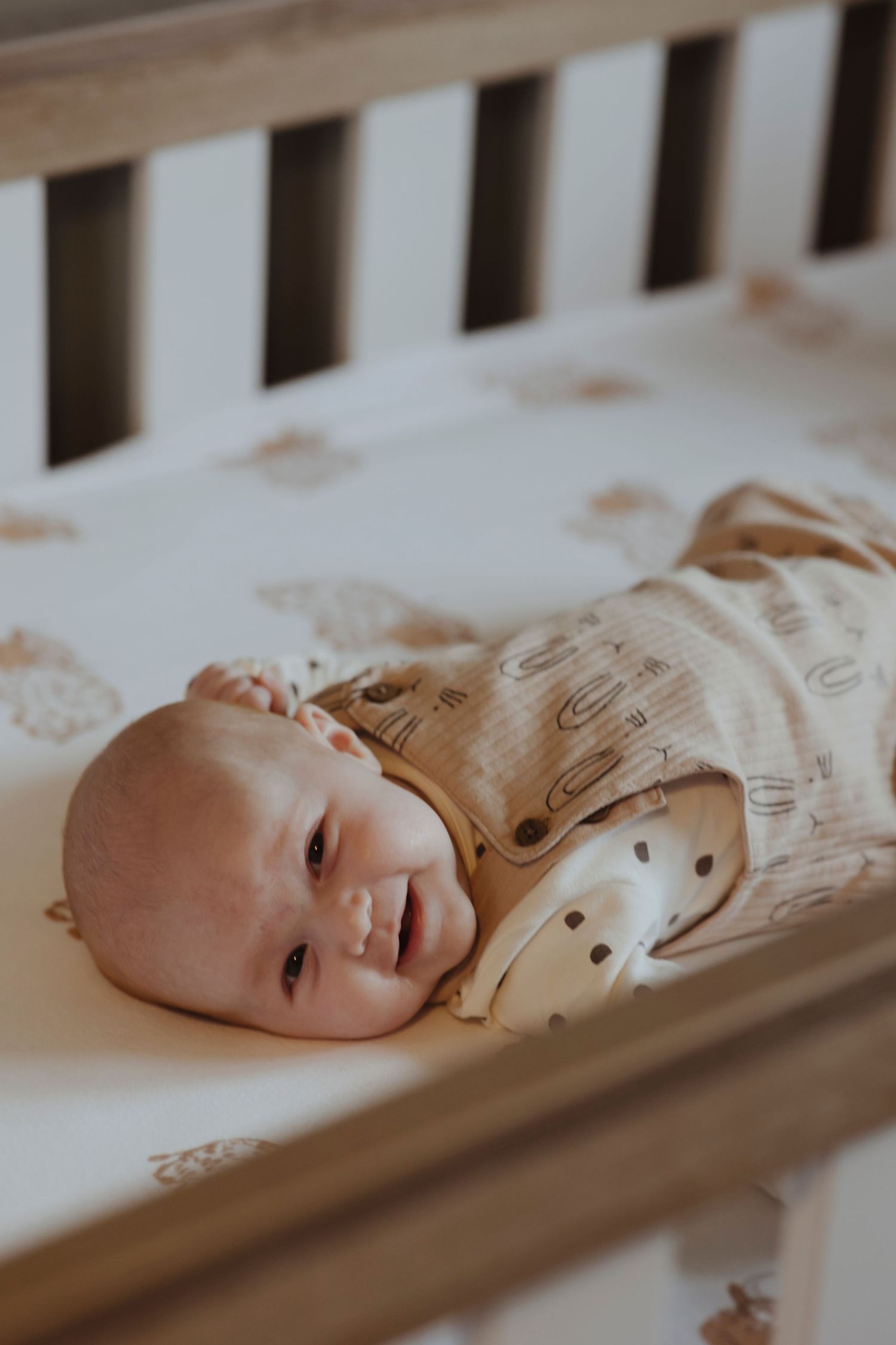 A baby lies in a crib, smiling softly while resting on a patterned sheet.