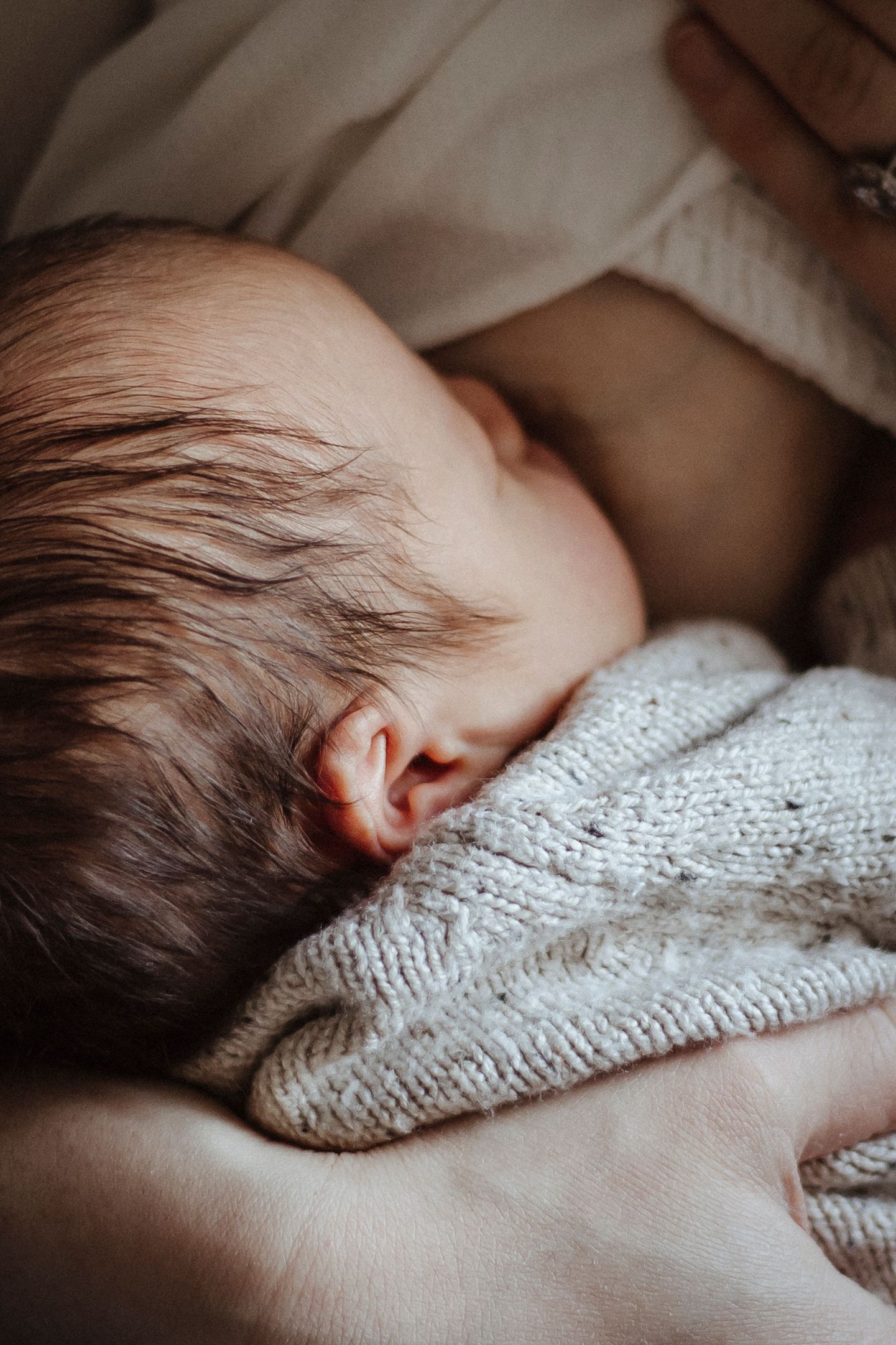A newborn nurses at a breast, held closely in a gentle embrace, wrapped in a soft knit outfit.