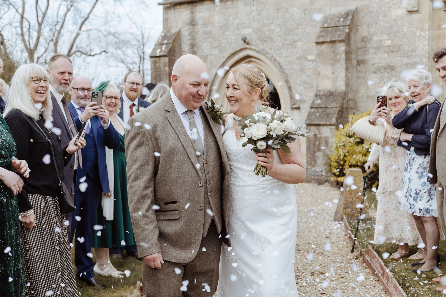 Newlyweds walk joyfully through guests tossing confetti as they smile warmly and hold a bouquet outside a stone building.