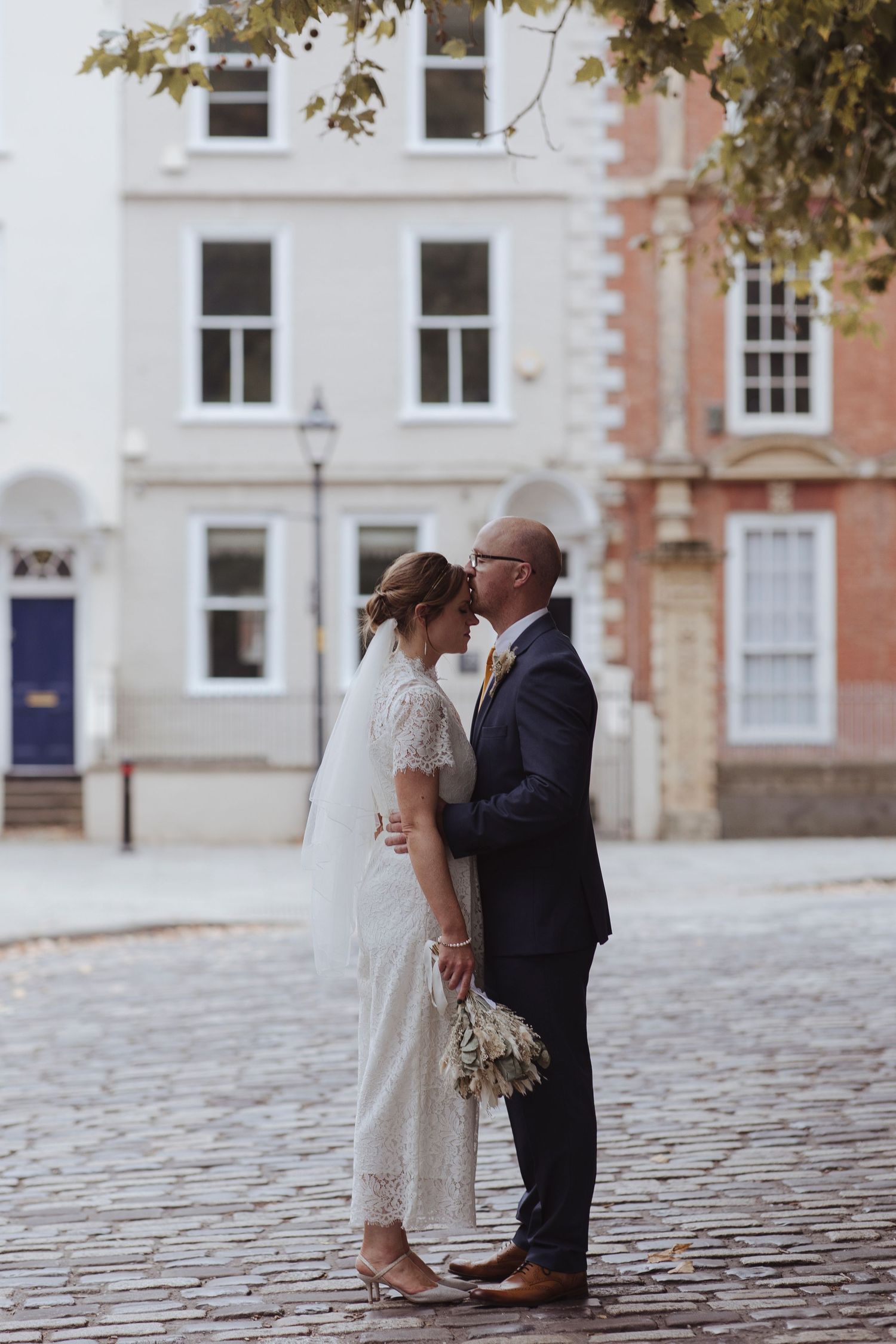 groom kissing bride on the forehead on cobbled streets with townhouses behind them