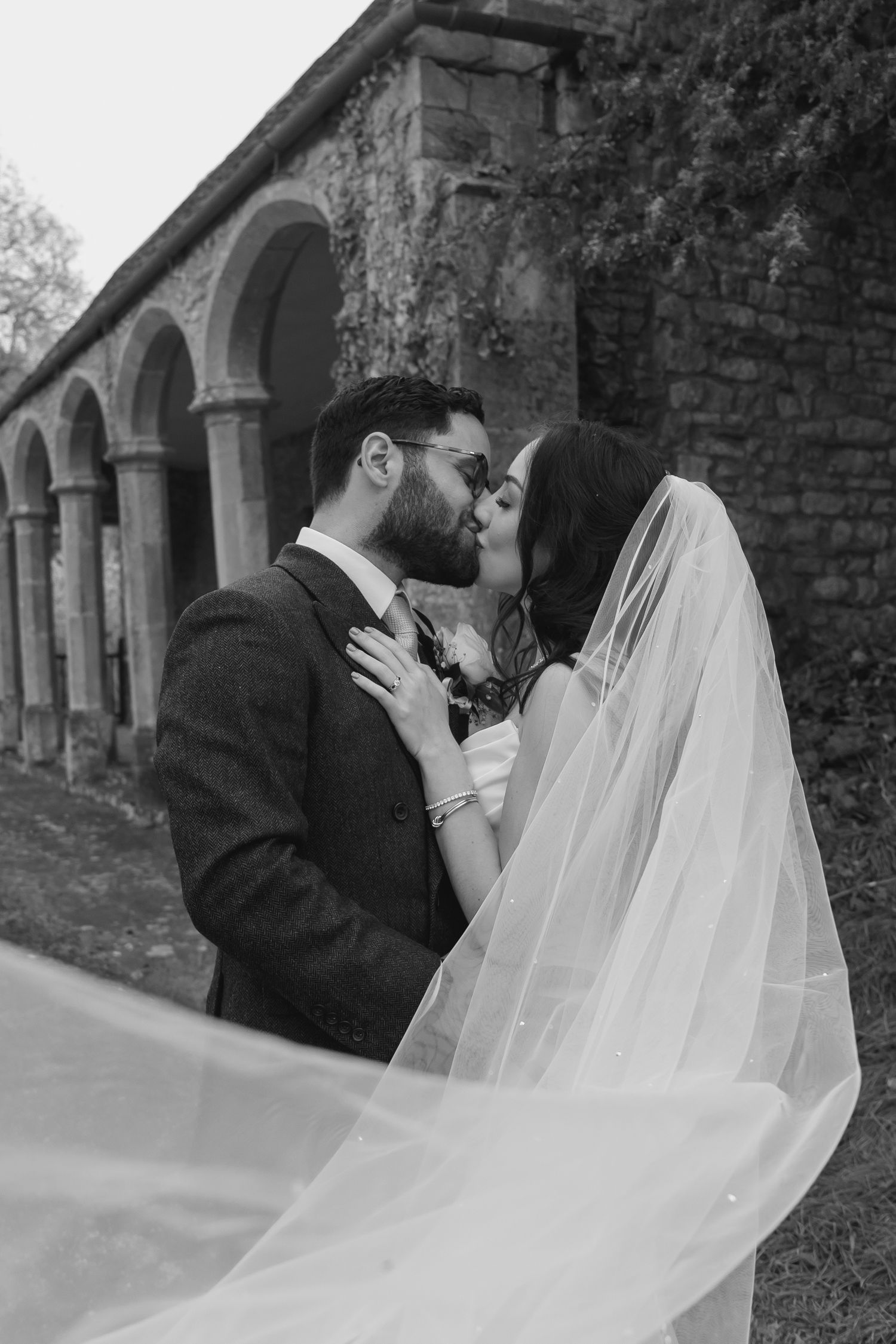 bride and groom kissing in front of stone arches whilst her veil sweeps across them