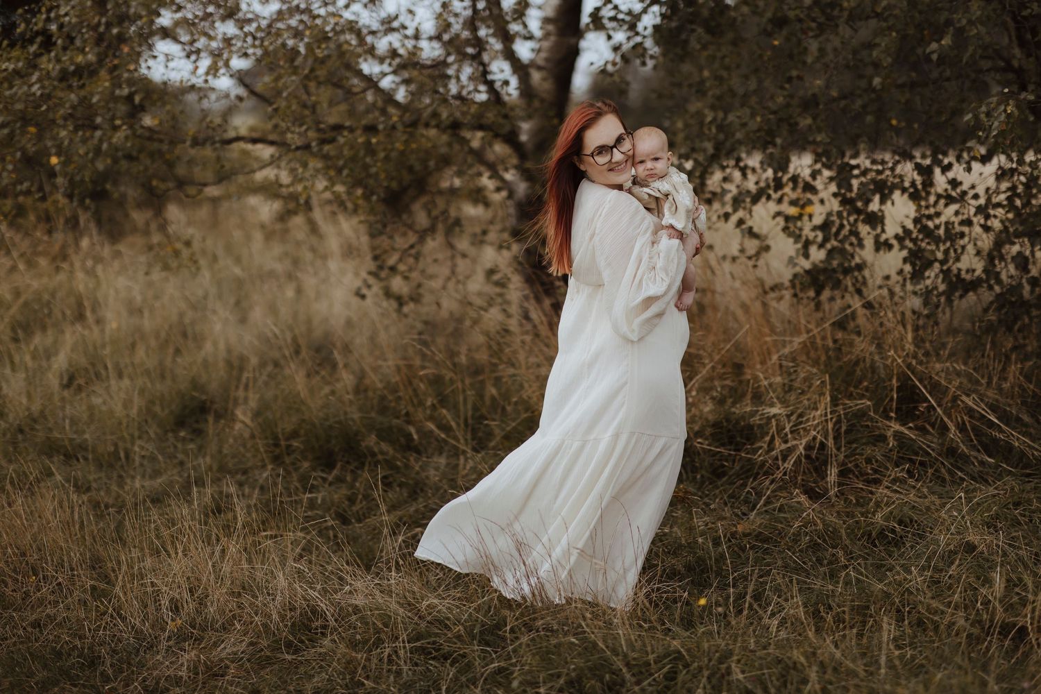 Mother holding her baby during an outdoor family photoshoot in a grassy field, wearing a flowing white dress, captured in warm natural light