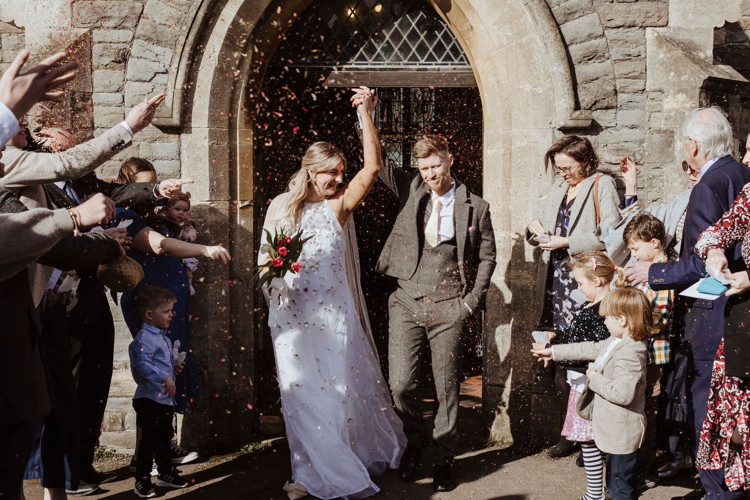 Joy spills into the sunlight as the newlyweds step out, showered in confetti and pure celebration.