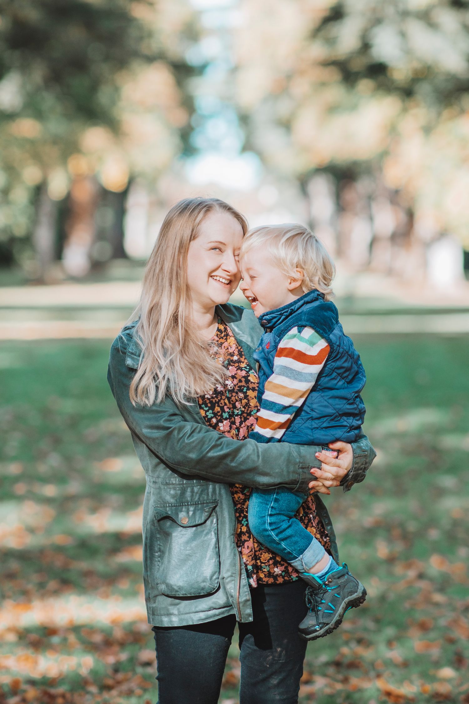 Mother holding a smiling child in an outdoor park, both looking at each other warmly amid trees and fallen leaves.