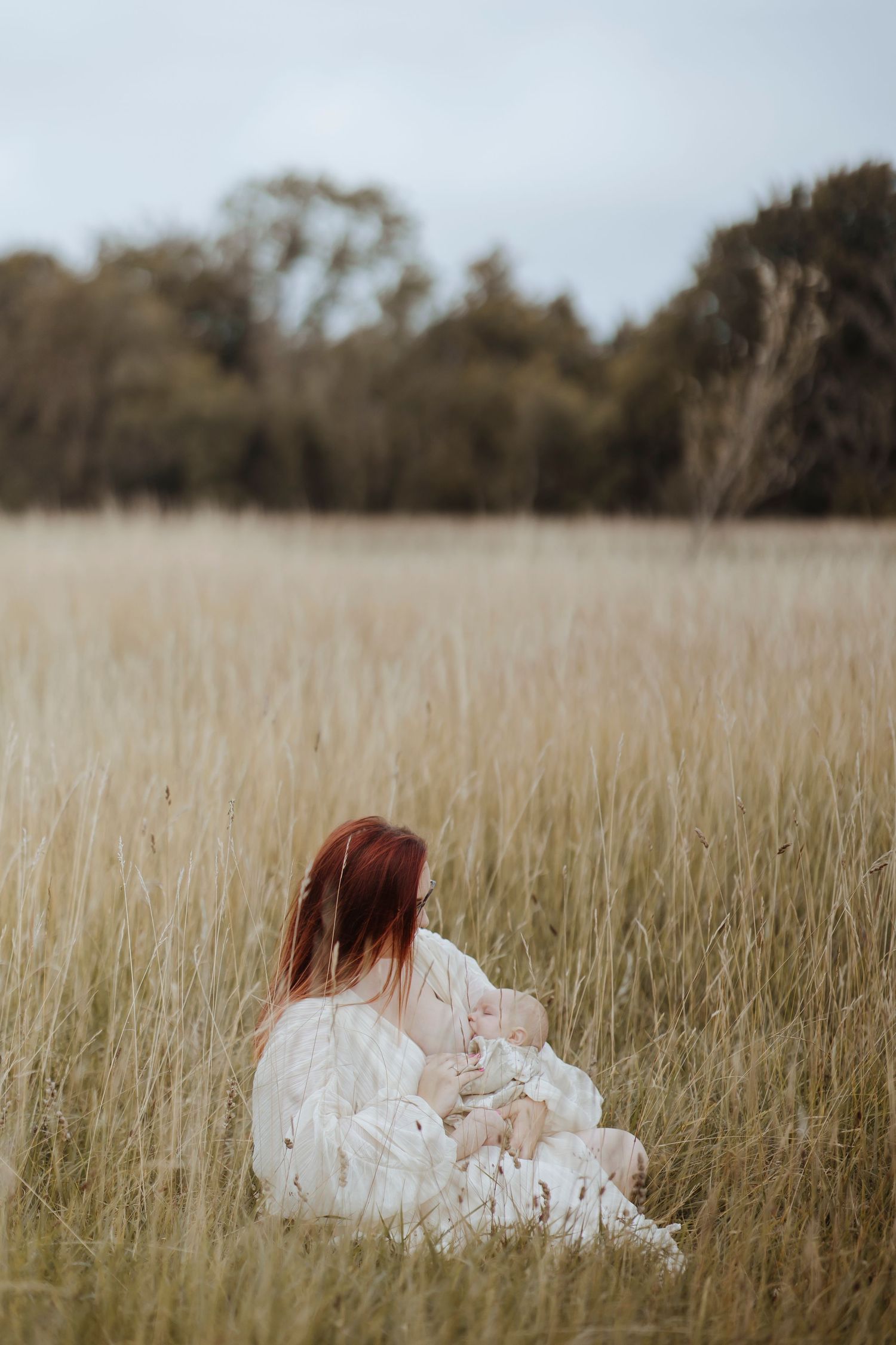 A woman with reddish hair sits in tall grass, holding a baby close in a peaceful, open field.