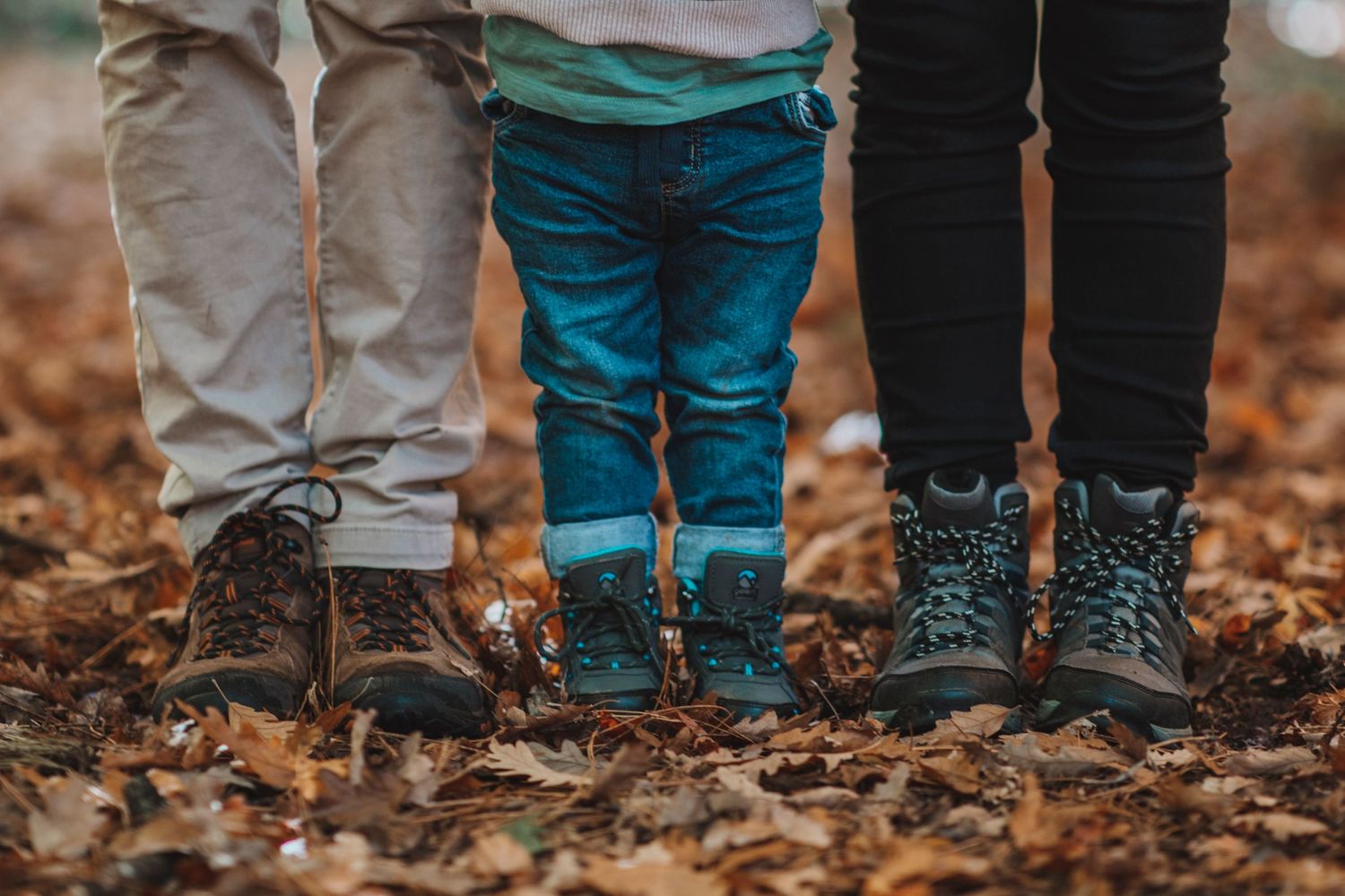 Close-up of two adults and a child standing together in autumn leaves, showing their boots and legs during an outdoor family walk.