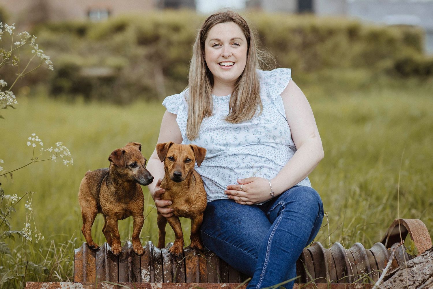 A woman sits outdoors with two small dogs beside her, enjoying a peaceful moment in a grassy field.