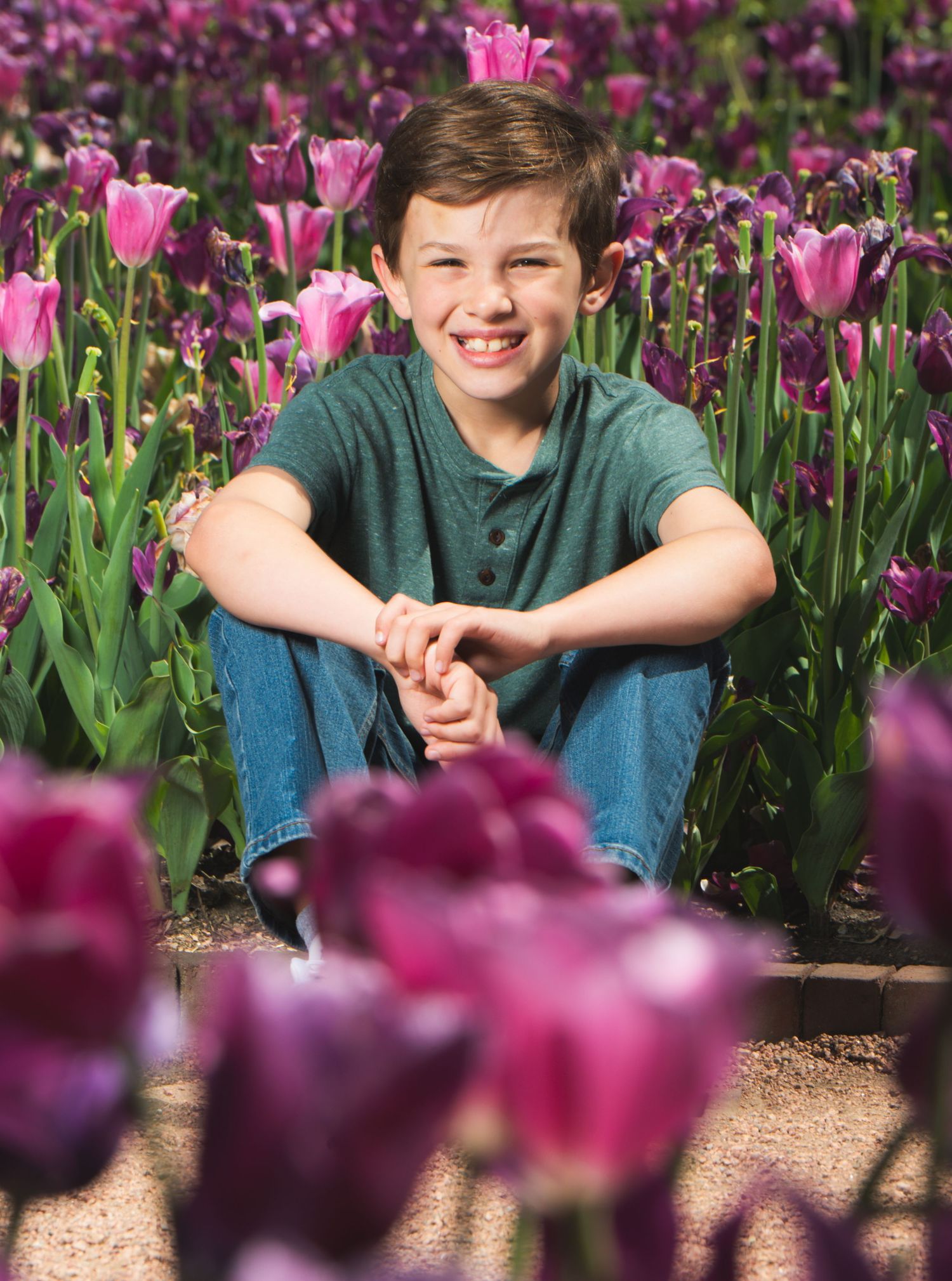 Smiling boy sitting among vibrant pink and purple tulips in a flower garden, wearing a teal shirt and jeans.