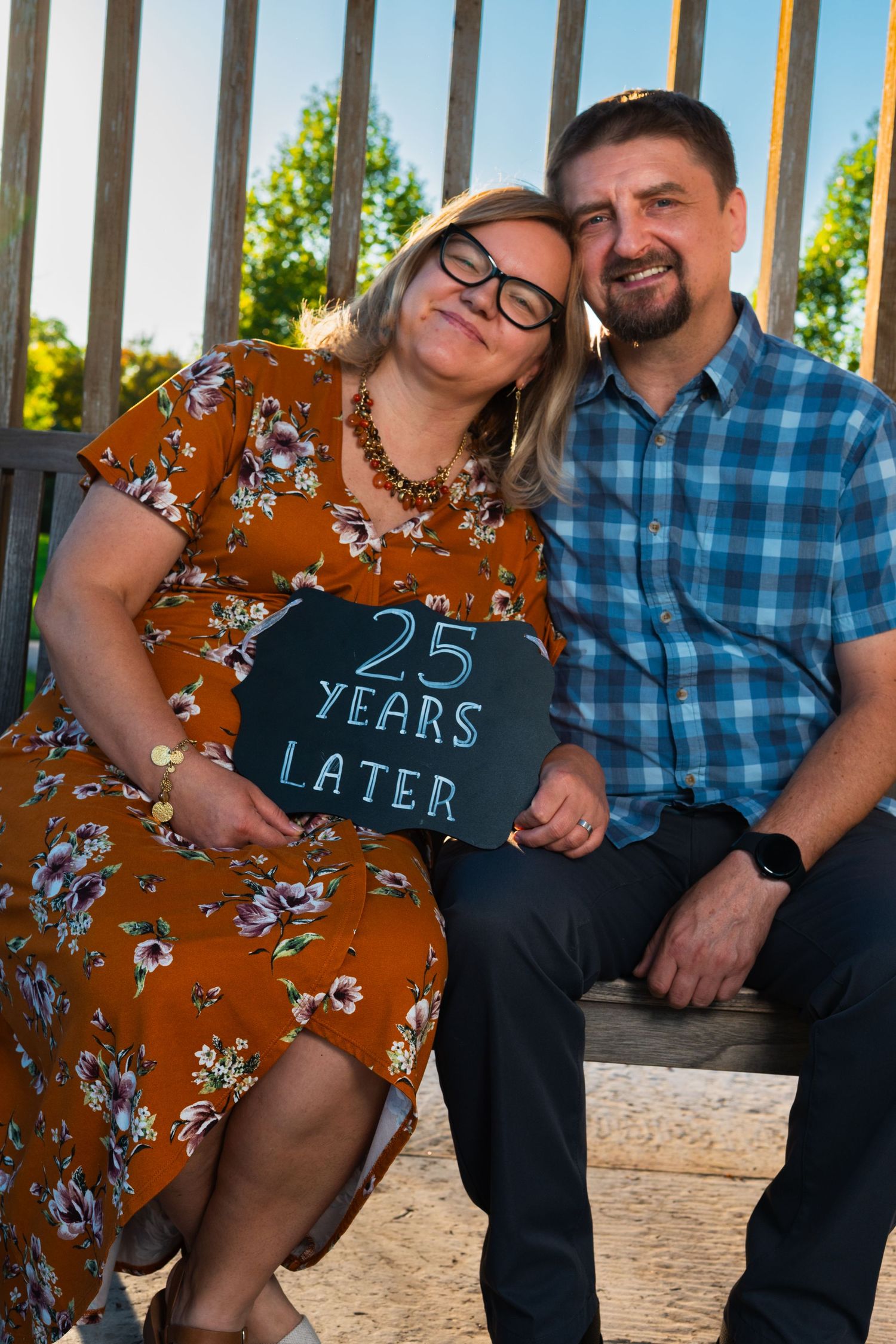 A smiling couple sits close together on a wooden bench holding a chalkboard sign that reads 25 Years Later.