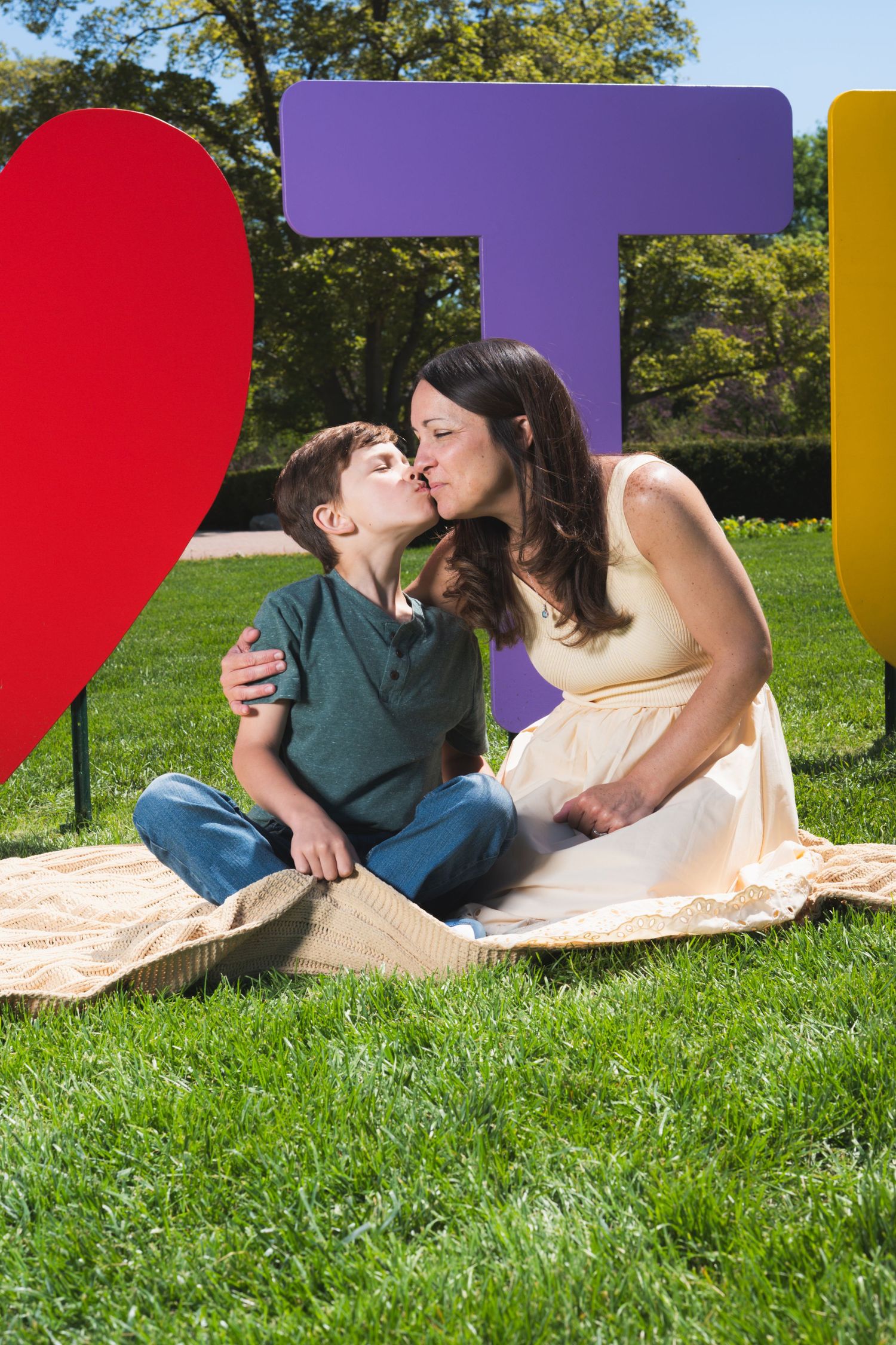 A boy kisses his pregnant mother on the lips while sitting on a blanket in a park with colorful letter decorations.