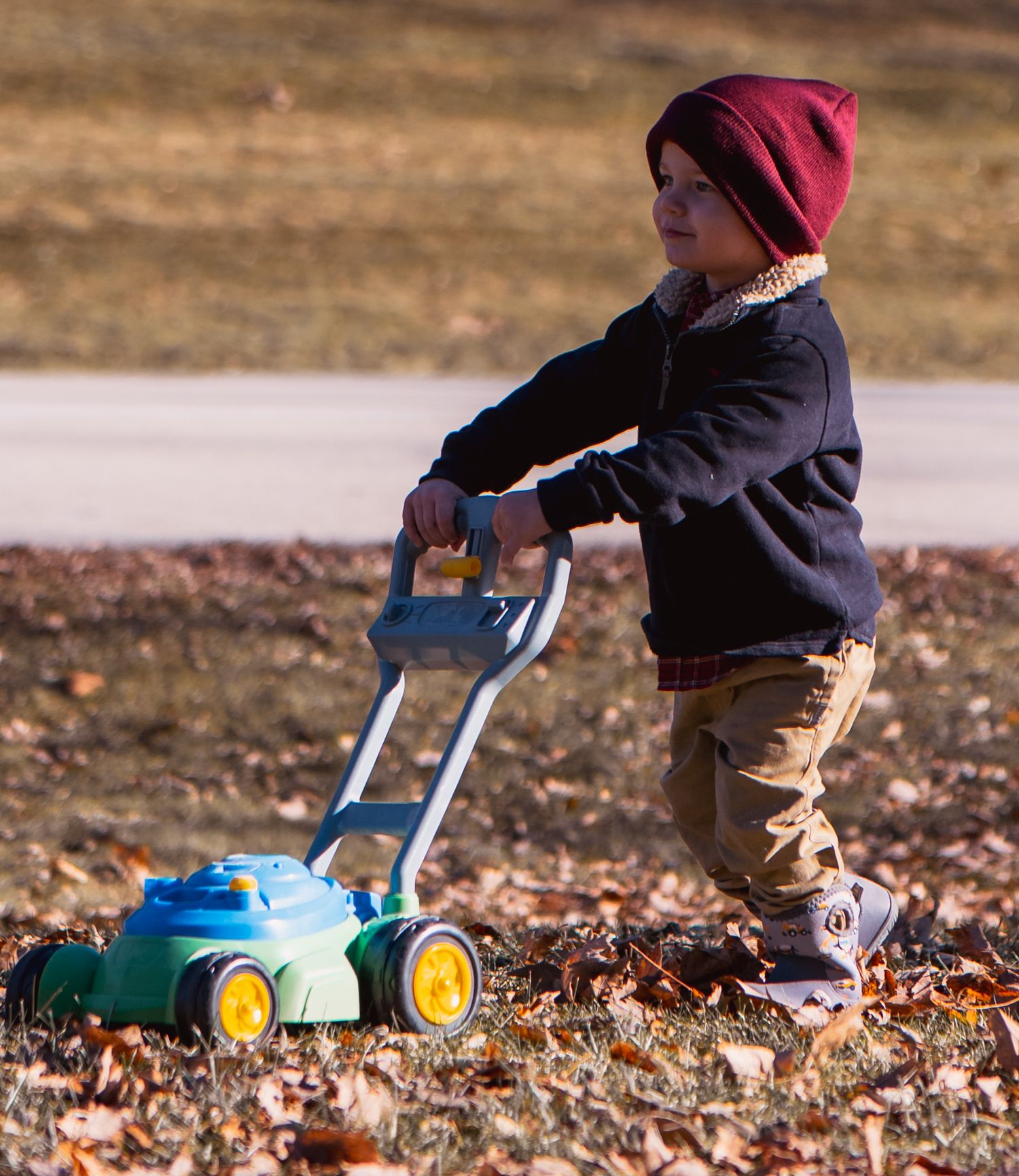 Small child in a burgundy beanie pushes a blue toy lawn mower through fallen autumn leaves.
