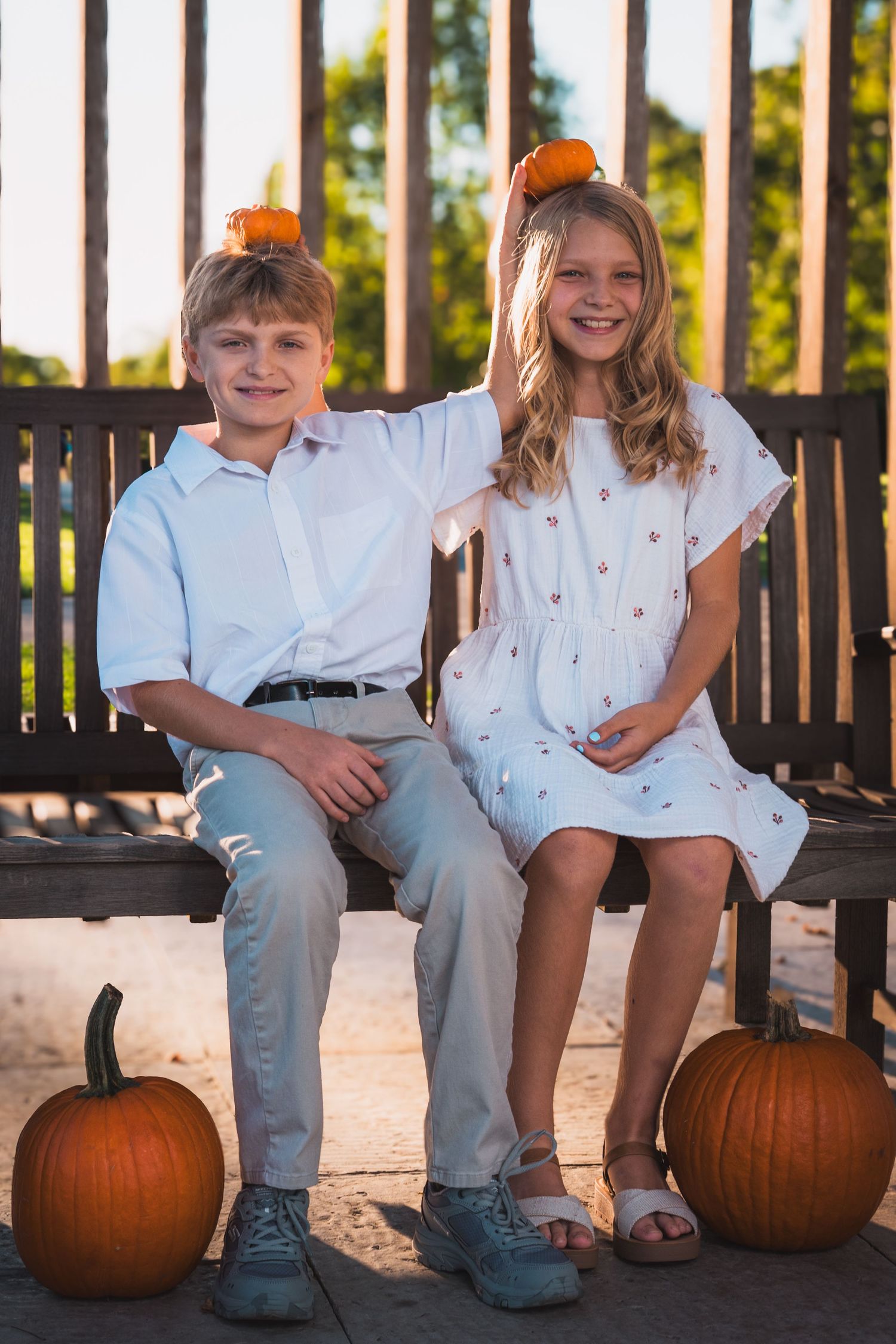 Smiling boy and girl sit on a wooden bench balancing small pumpkins on their heads, with larger pumpkins on the ground beside them.