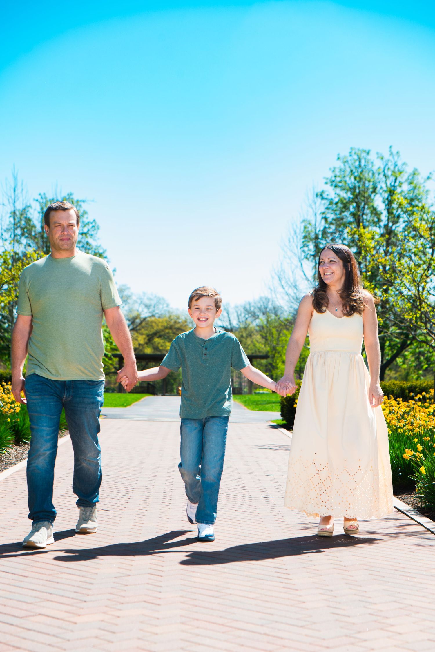Happy family of three holding hands walking on a brick path in a sunny park with blue sky and blooming trees.