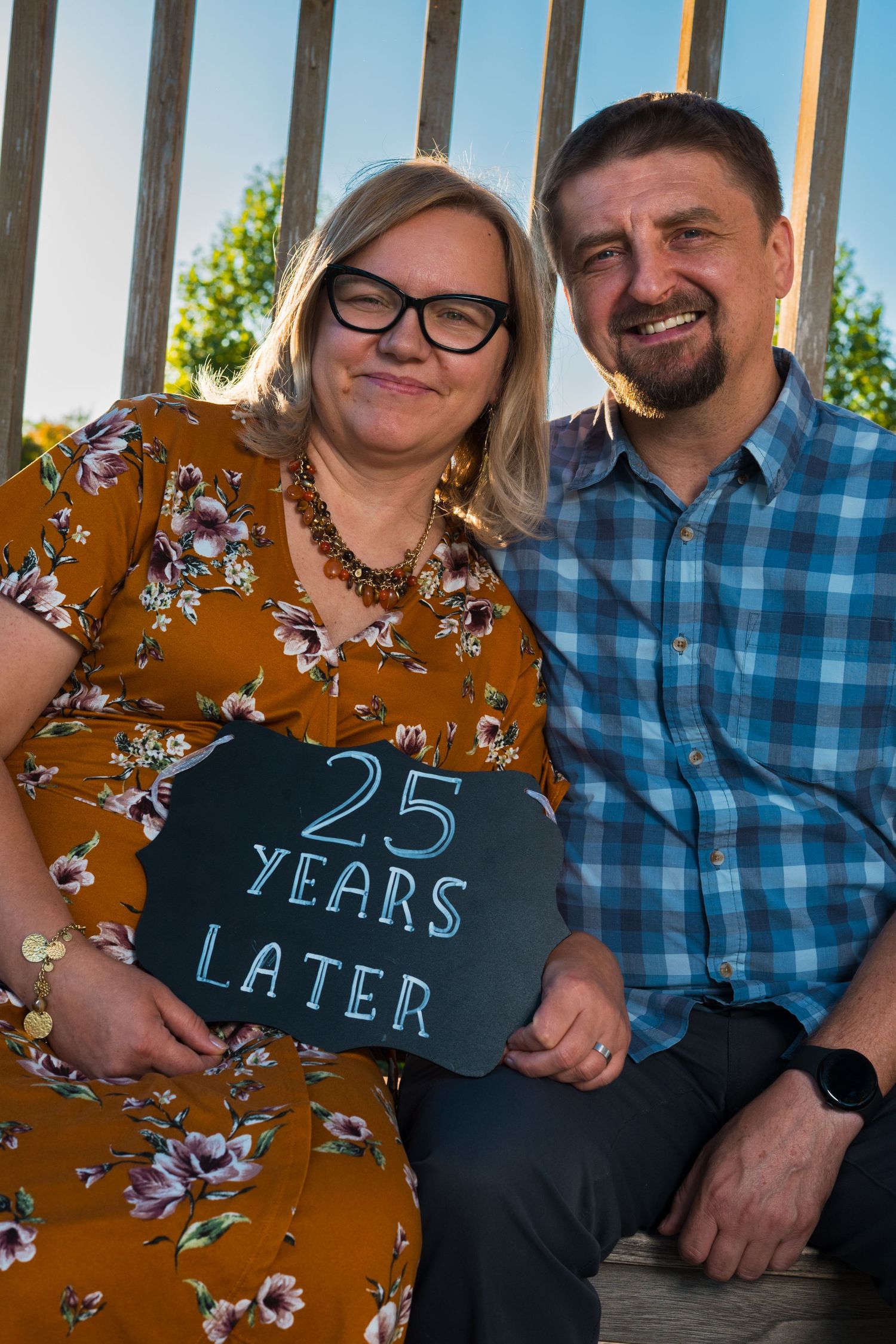 Smiling couple sitting together on a wooden bench outdoors, holding a chalkboard sign that reads 25 Years Later.
