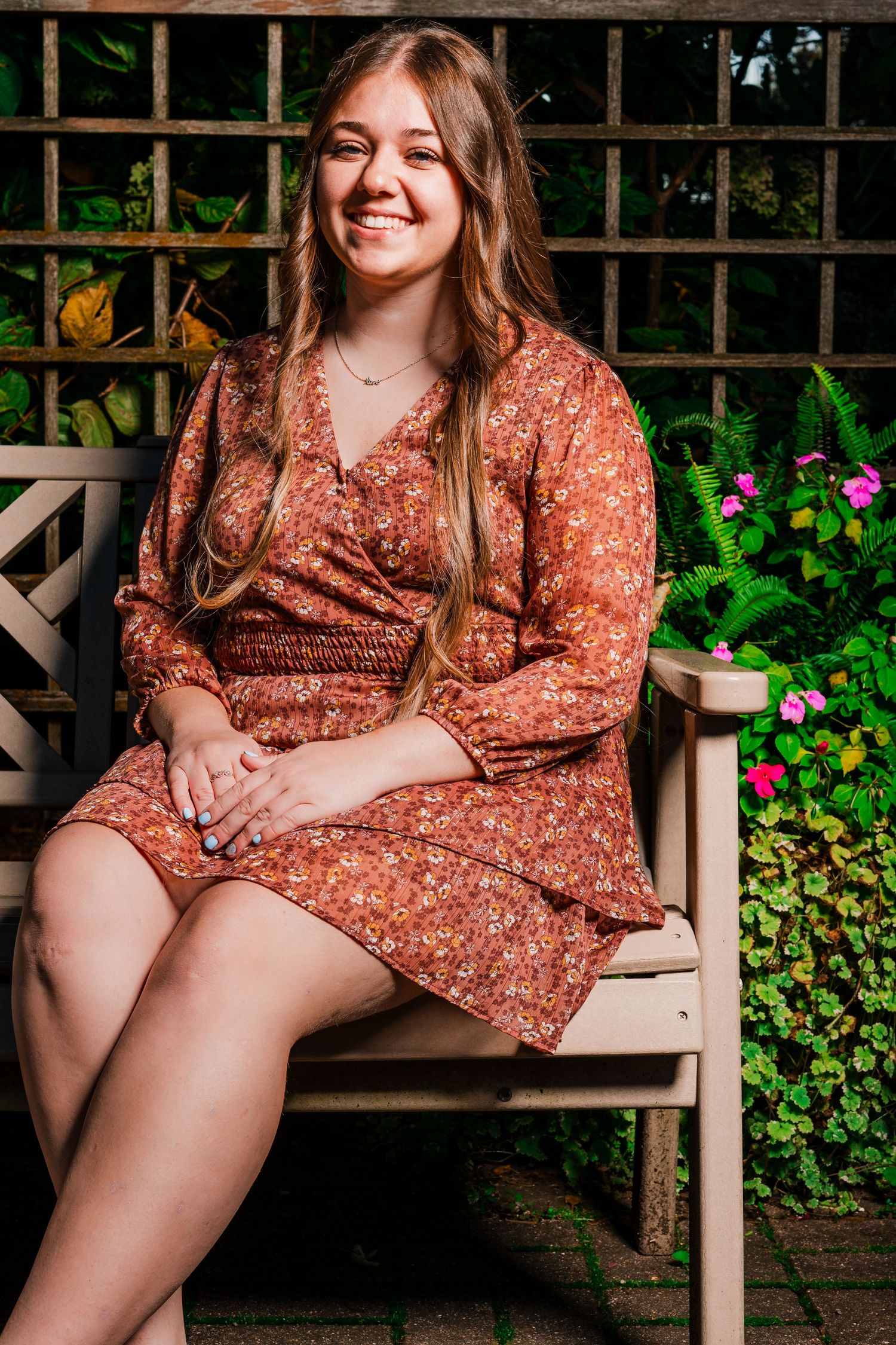 Smiling young woman with long hair in a rust floral wrap dress sits on a wooden garden bench beside pink flowers and green plants.