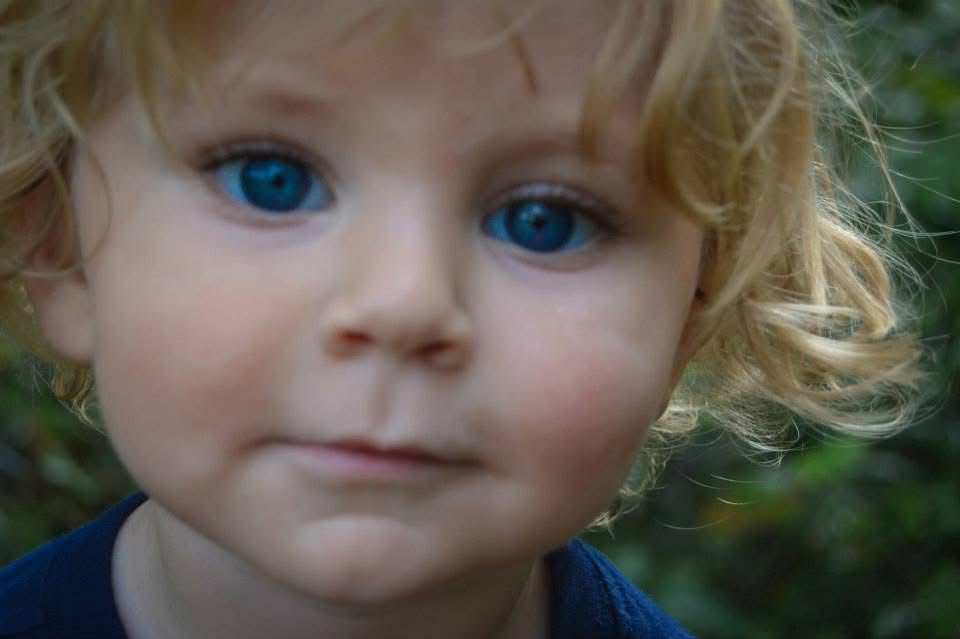 A toddler with blonde curly hair and bright blue eyes smiles sweetly at the camera in an outdoor setting.
