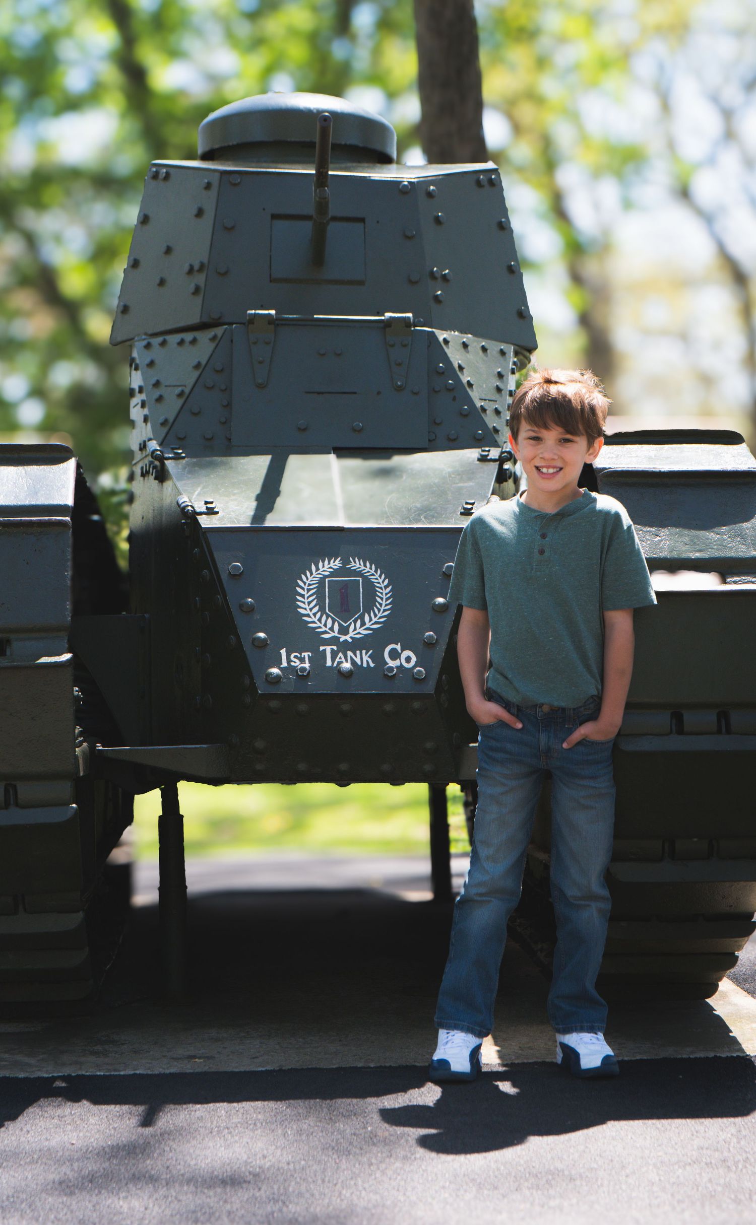Young boy smiling beside a vintage 1st Tank Co. military tank display outdoors on a sunny day.