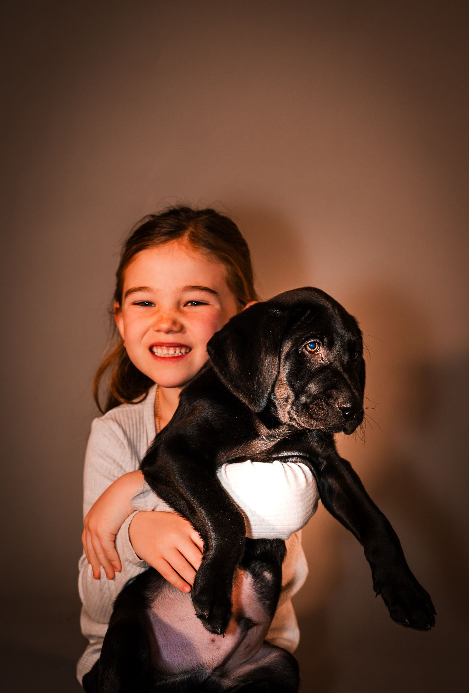 A young child holds a black puppy while smiling against a warm brown background.