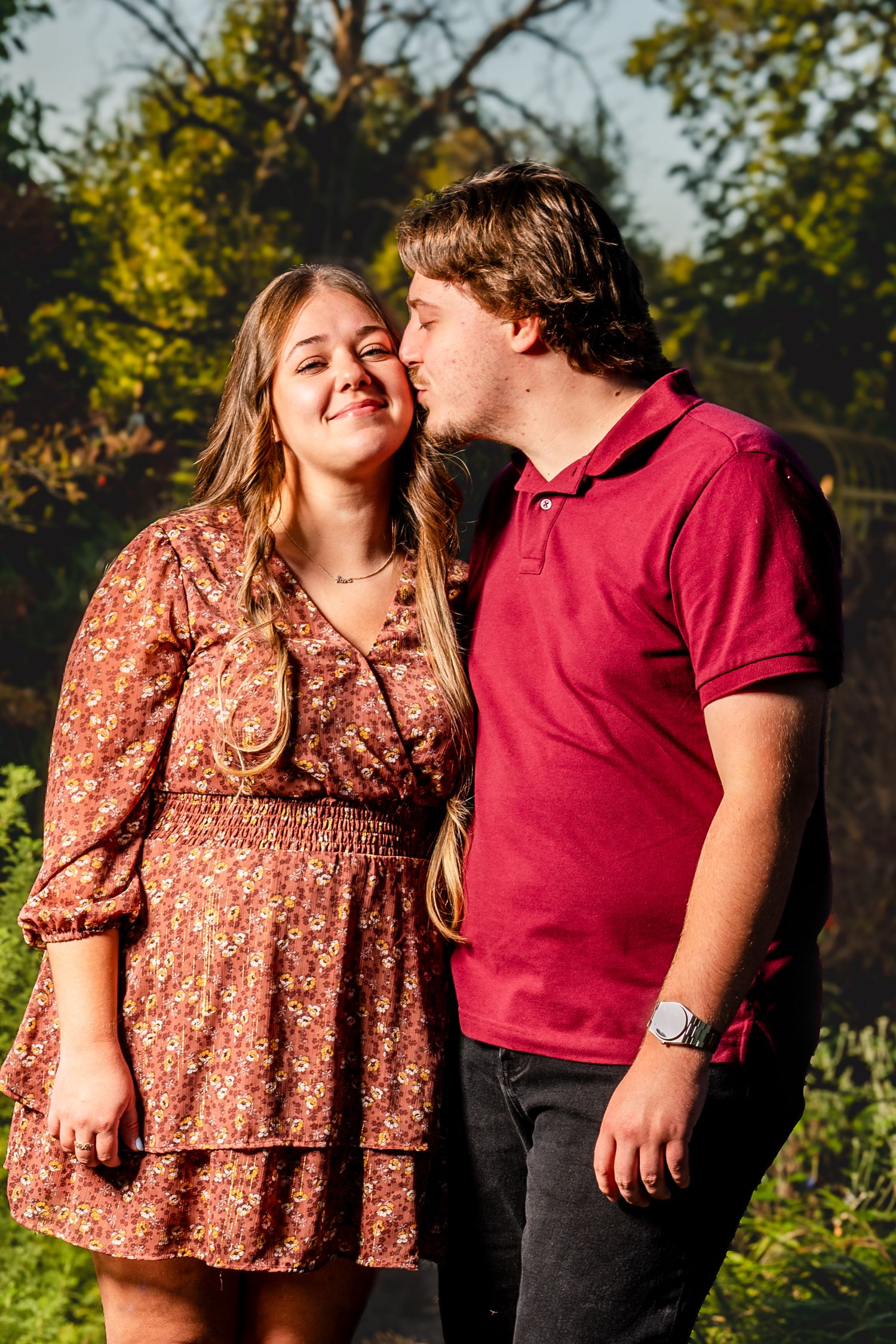 Young man in a red polo kisses the cheek of a smiling woman in a floral dress during an outdoor couple portrait surrounded by green trees.