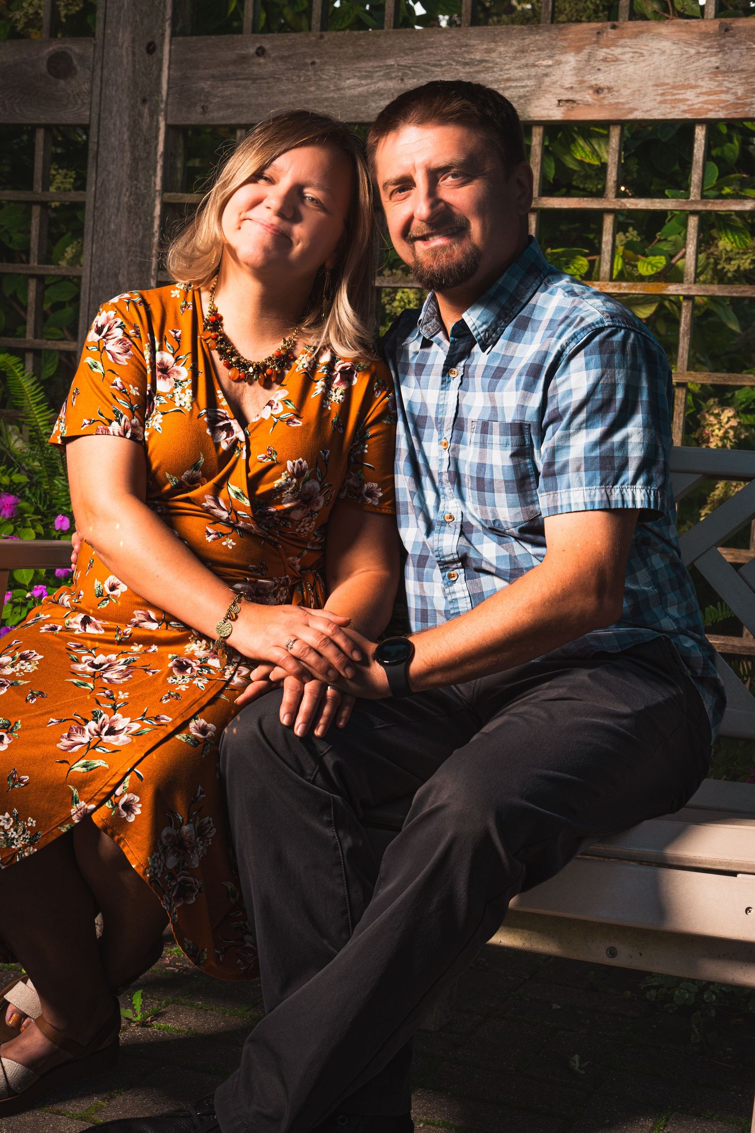 Smiling couple in floral dress and plaid shirt sitting close on a garden bench, holding hands in warm evening sunlight.