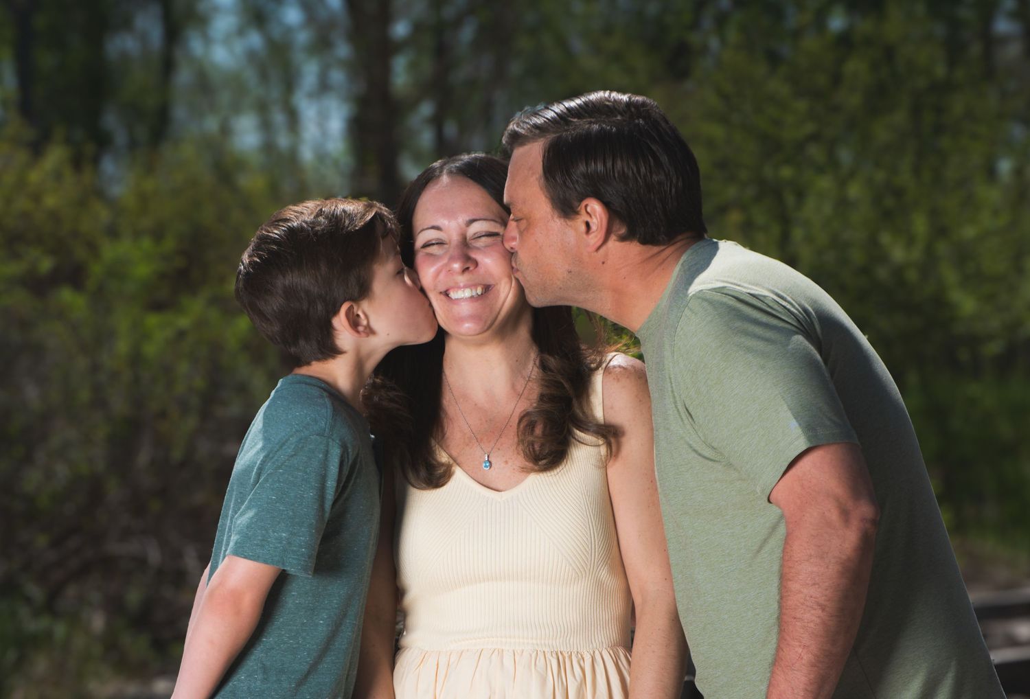 A smiling woman receives kisses on both cheeks from a young boy and a man outdoors with trees in the background.