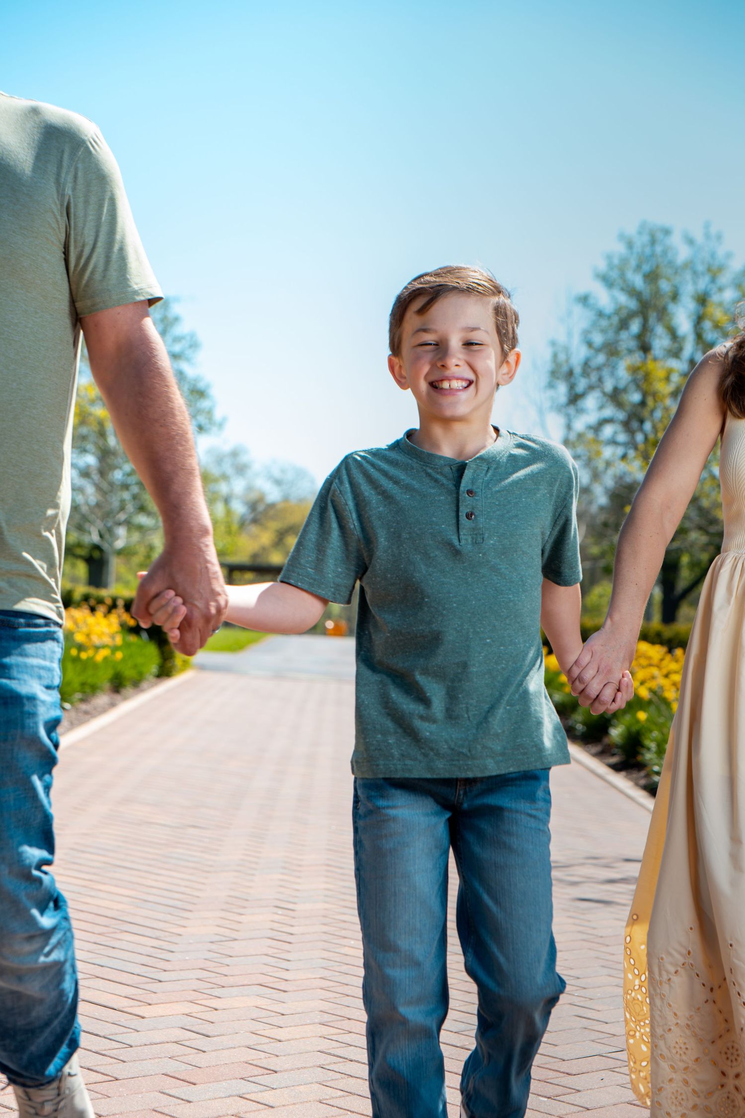 Happy boy holds hands with parents walking on brick path in sunny park with yellow flowers and blue sky.