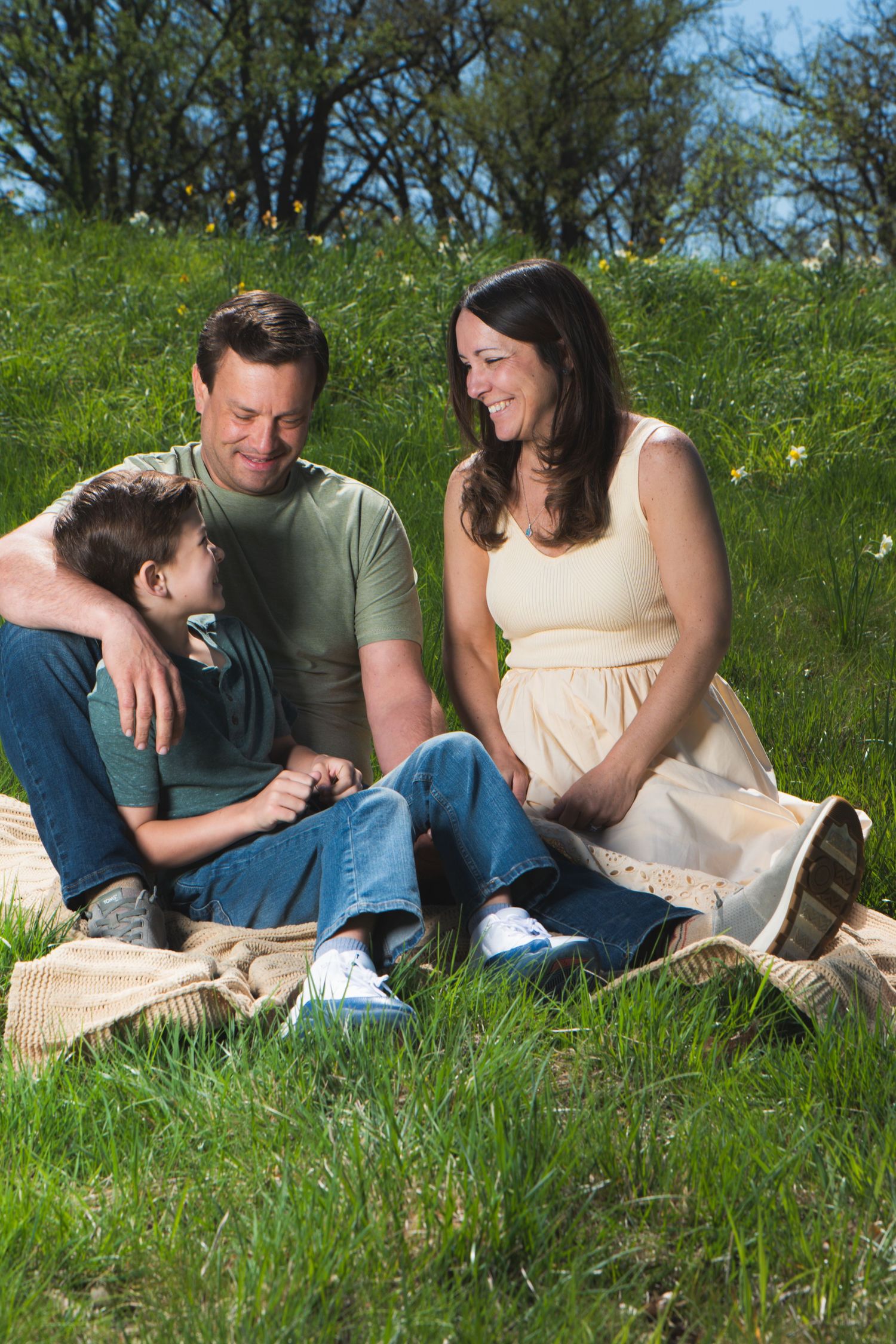 A happy family sitting on a blanket in a green meadow, with a pregnant mother, father, and young child smiling together.