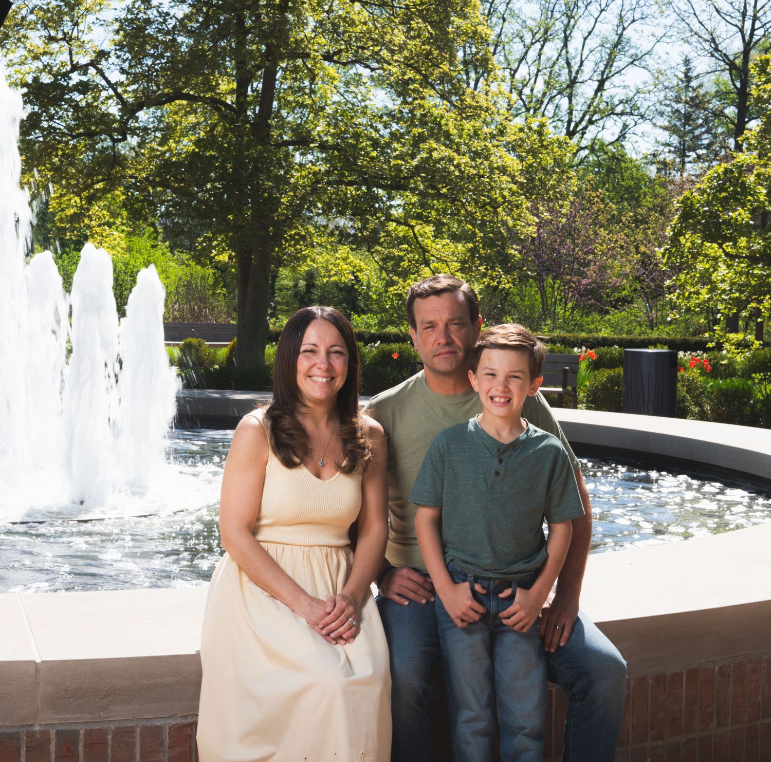 Family of three smiling together in front of a decorative fountain in a lush green park on a sunny day.
