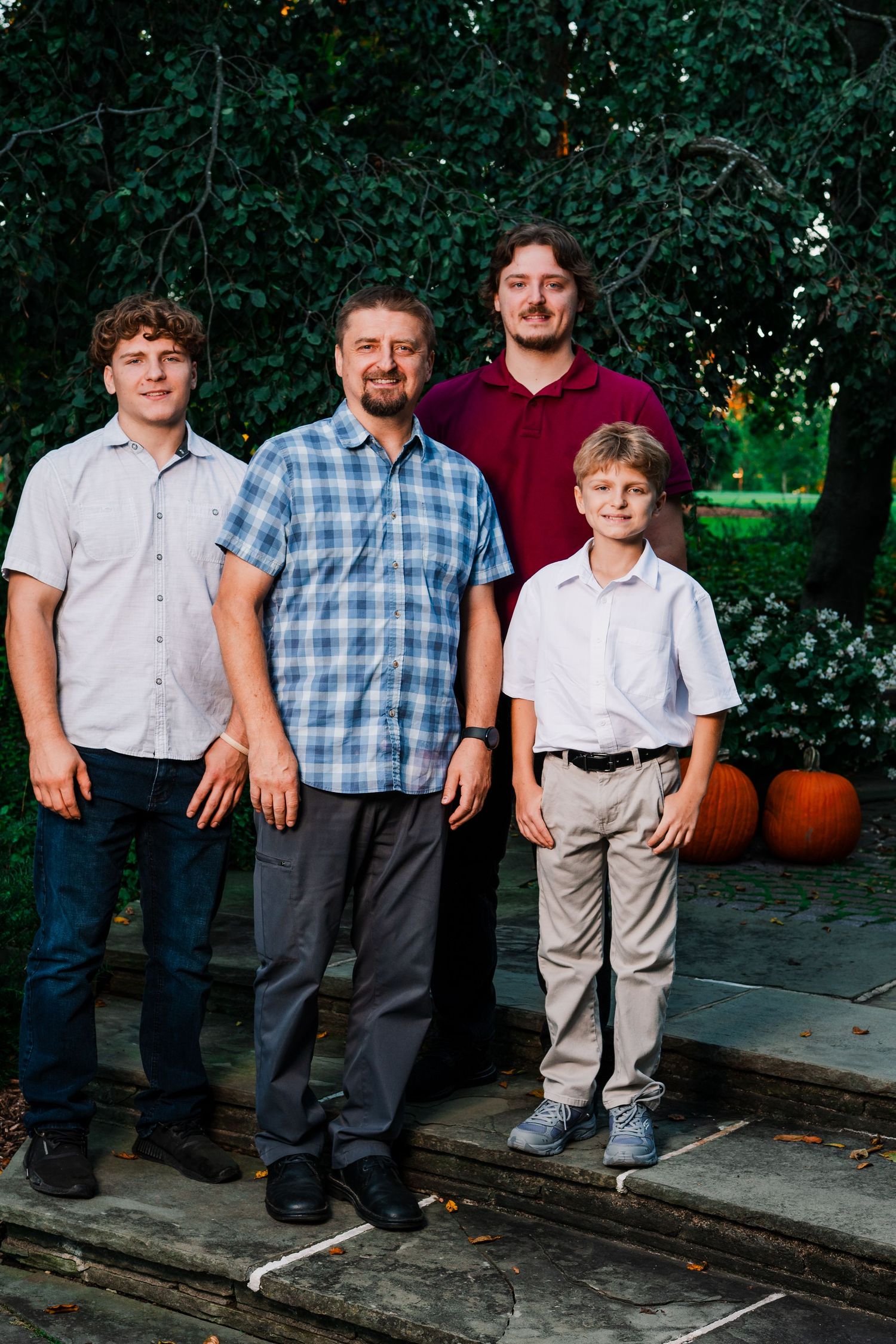 A man stands with three younger males on stone steps outdoors near green trees and orange pumpkins in a family portrait.
