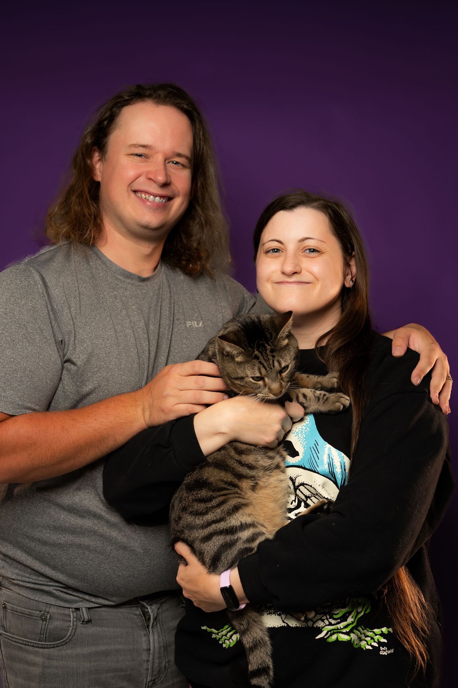 A smiling couple holds a tabby kitten against a purple background, posing for a portrait photo together.