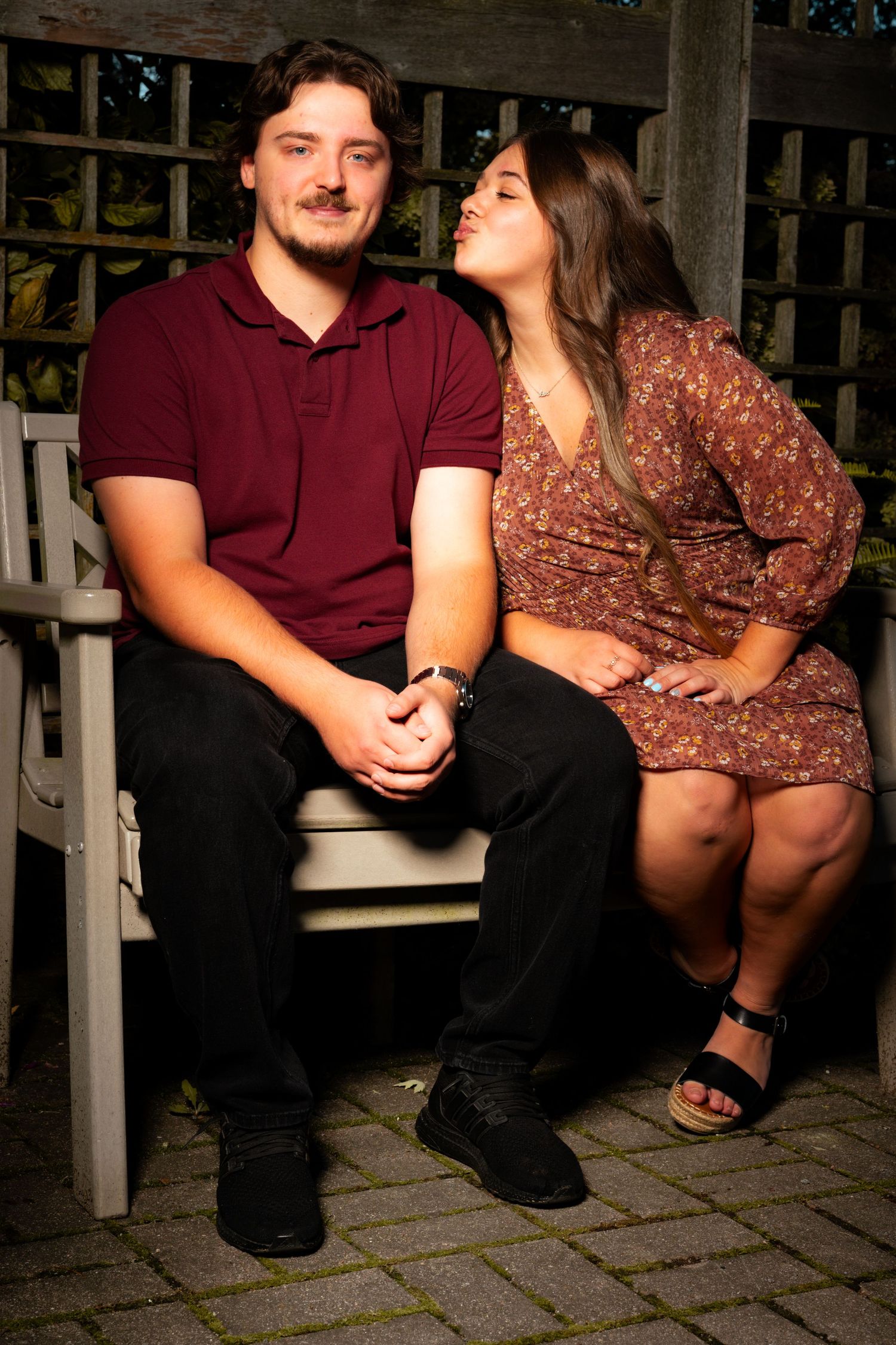 A young couple sits close together on a wooden garden bench while the woman leans in to give the man a kiss on the cheek.