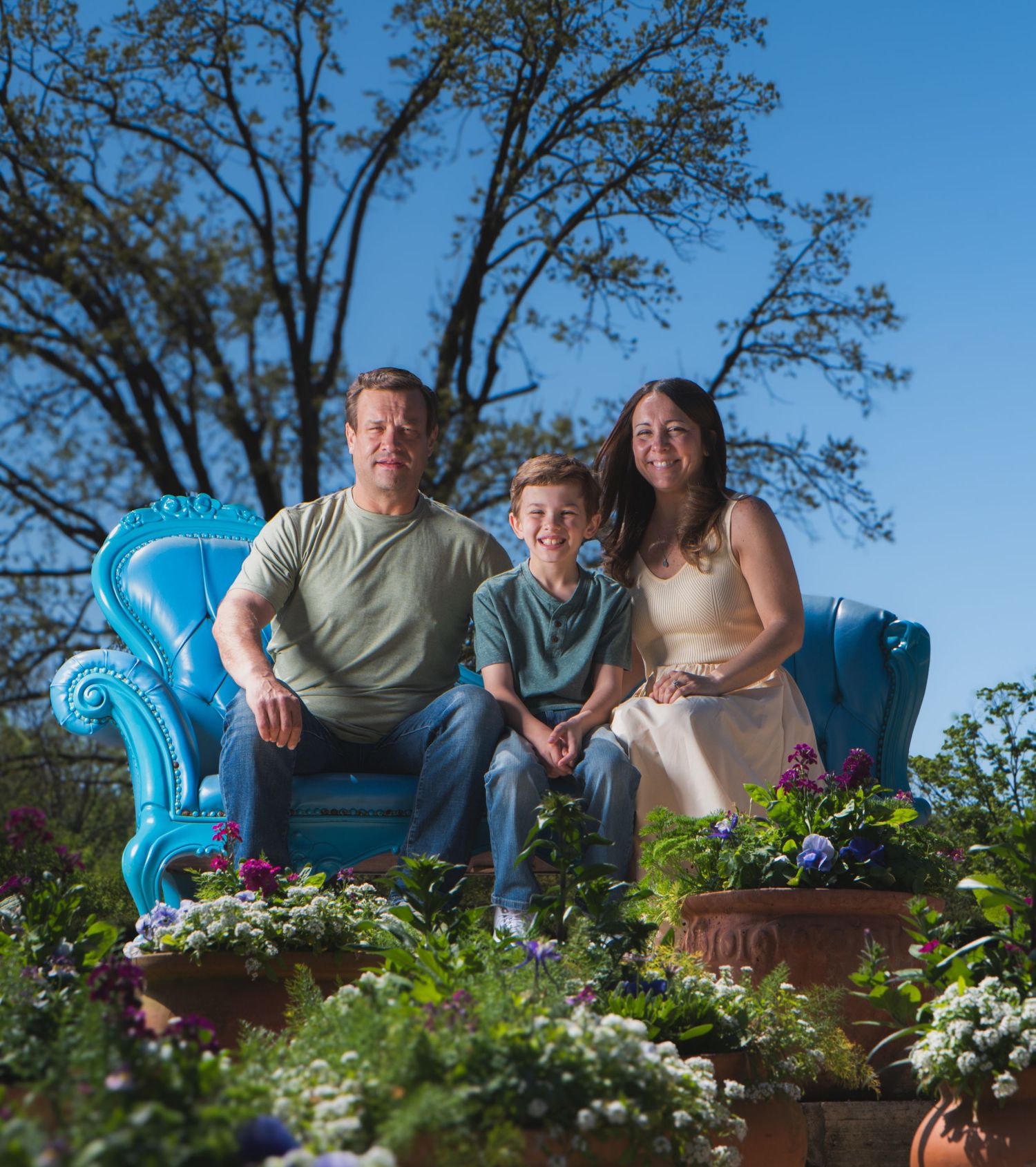 Family of three sitting on a blue armchair surrounded by colorful flowers and terracotta pots outdoors.