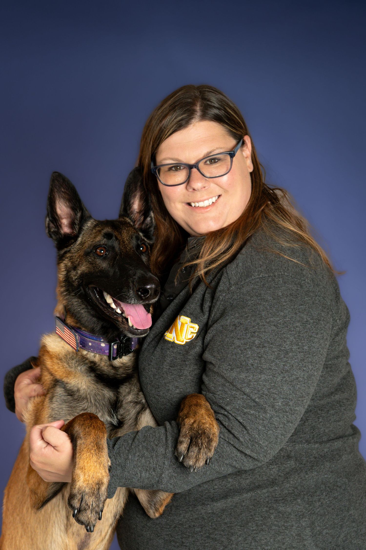 Woman with glasses hugging a Belgian Malinois dog, smiling for a studio portrait photo on blue background.