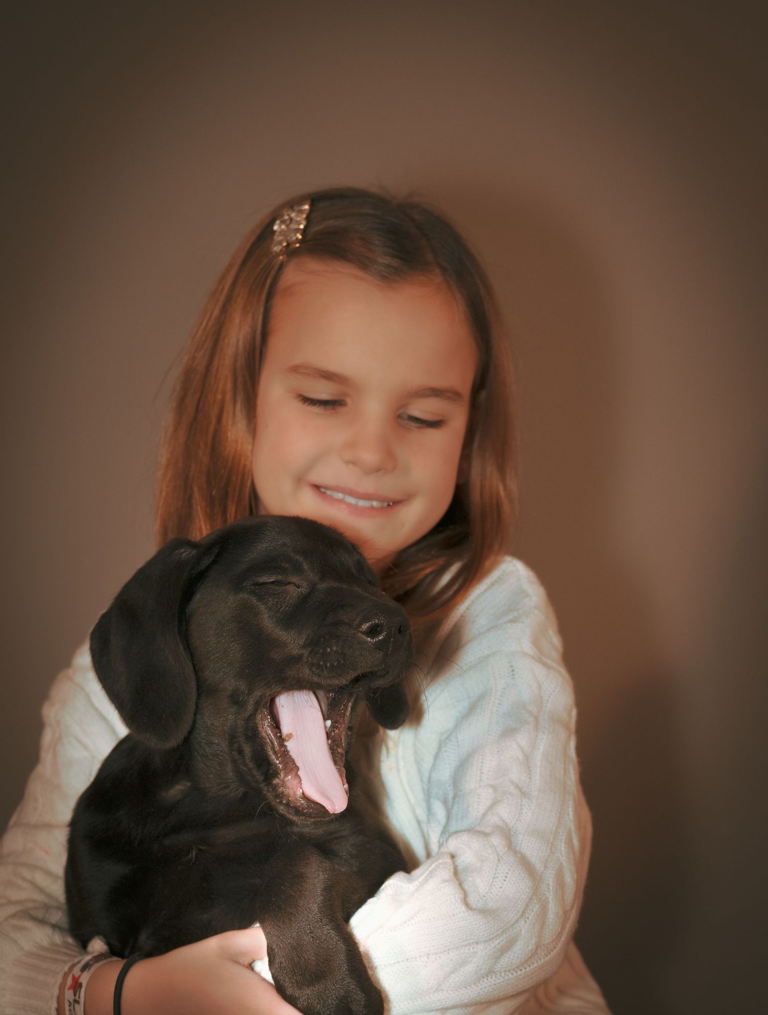 A young person in a white sweater hugs and cuddles with a yawning black Labrador puppy.