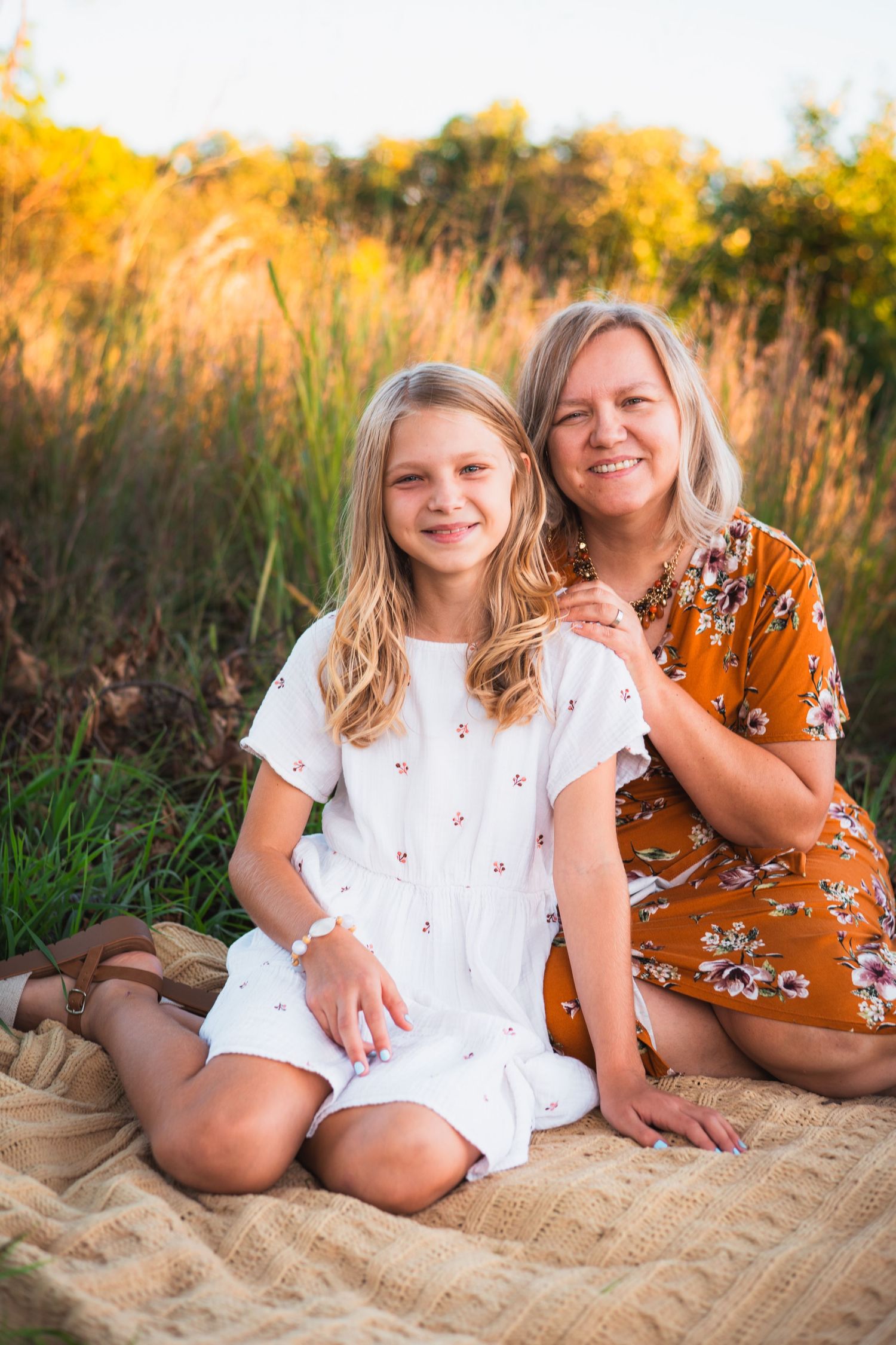 Smiling girl in a white dress sits on a blanket beside a woman in an orange floral dress in front of golden autumn grass.