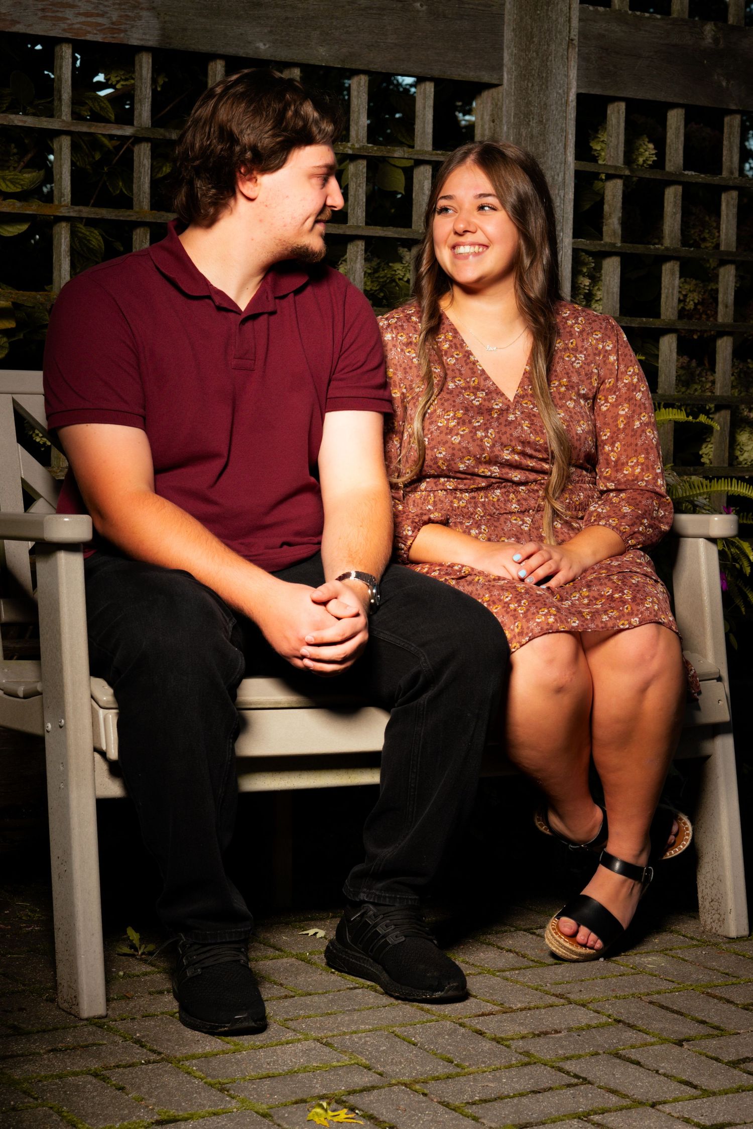 A smiling young couple sits close together on a wooden garden bench, looking at each other in a cozy outdoor portrait setting.