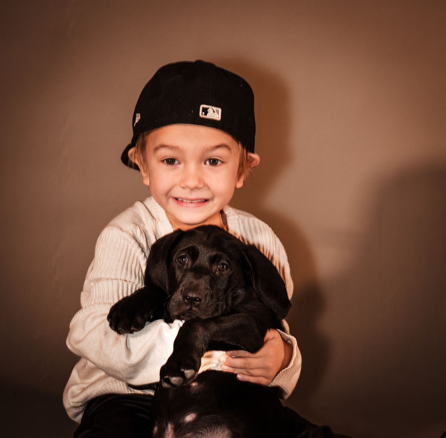 A series of photos showing a child in a baseball cap posing with a small black puppy against a brown background.