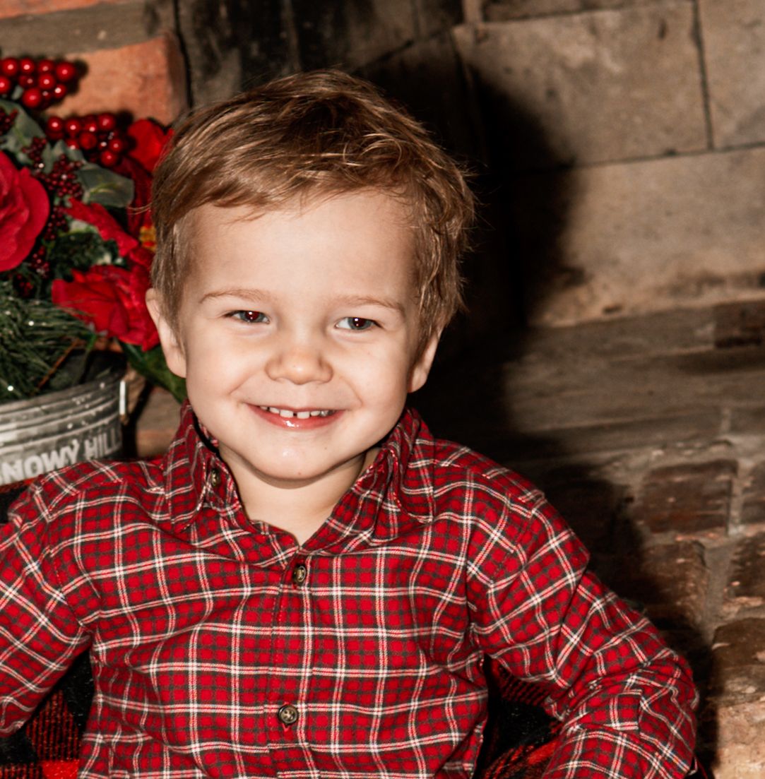 A young child in a red plaid shirt smiles brightly during a holiday photo session with poinsettias in the background.