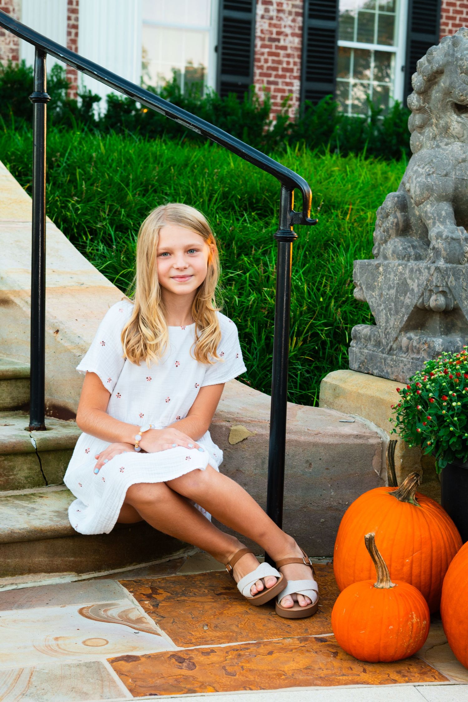 Smiling girl in a white dress and sandals sits on stone steps beside pumpkins, a mum plant, and a stone lion statue.