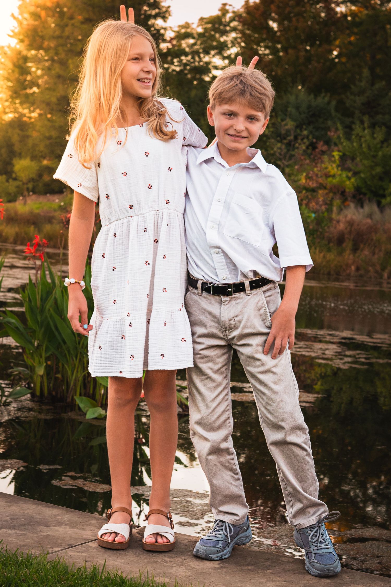 Two smiling children stand side by side playfully making bunny ears by a pond at sunset, wearing white outfits and sandals.