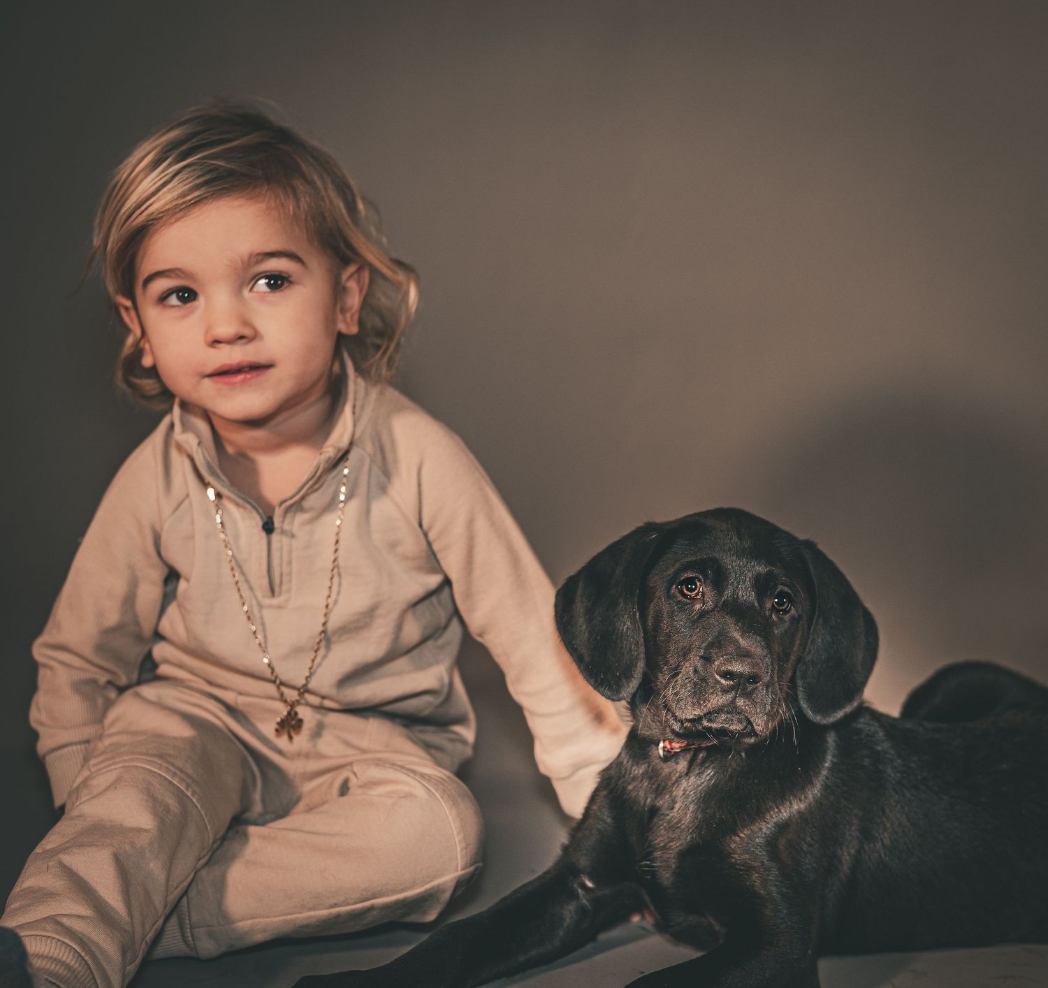 Child in beige sweater sitting on couch with black dog and gray cat in moody lighting series.