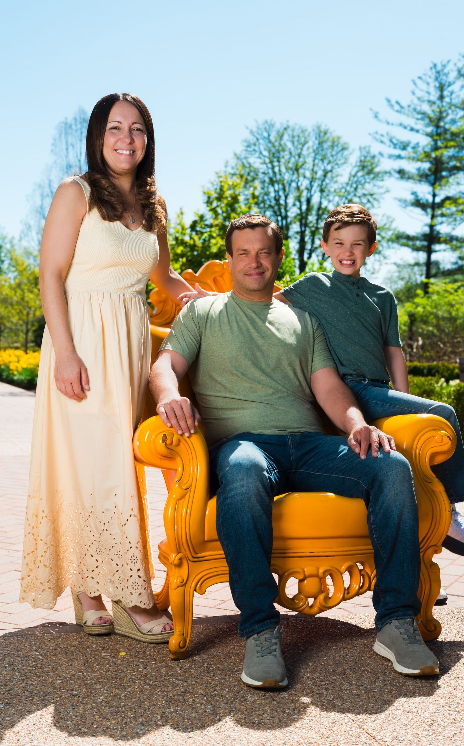 Family of three posing outdoors on sunny day with ornate yellow chair, woman in cream dress, man in green shirt, boy standing behind.