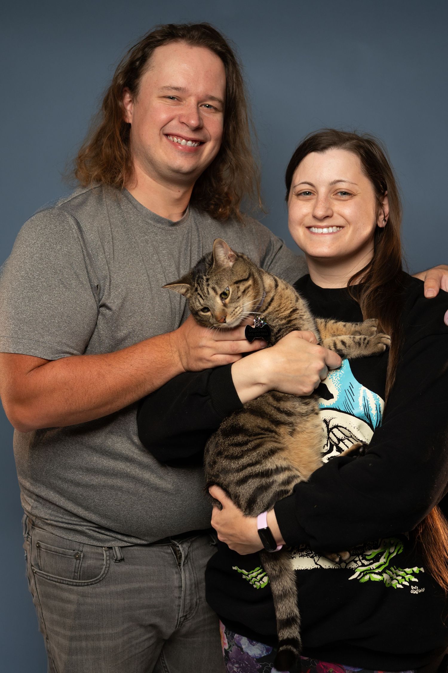 A smiling couple holds a tabby cat together against a blue-gray studio backdrop.